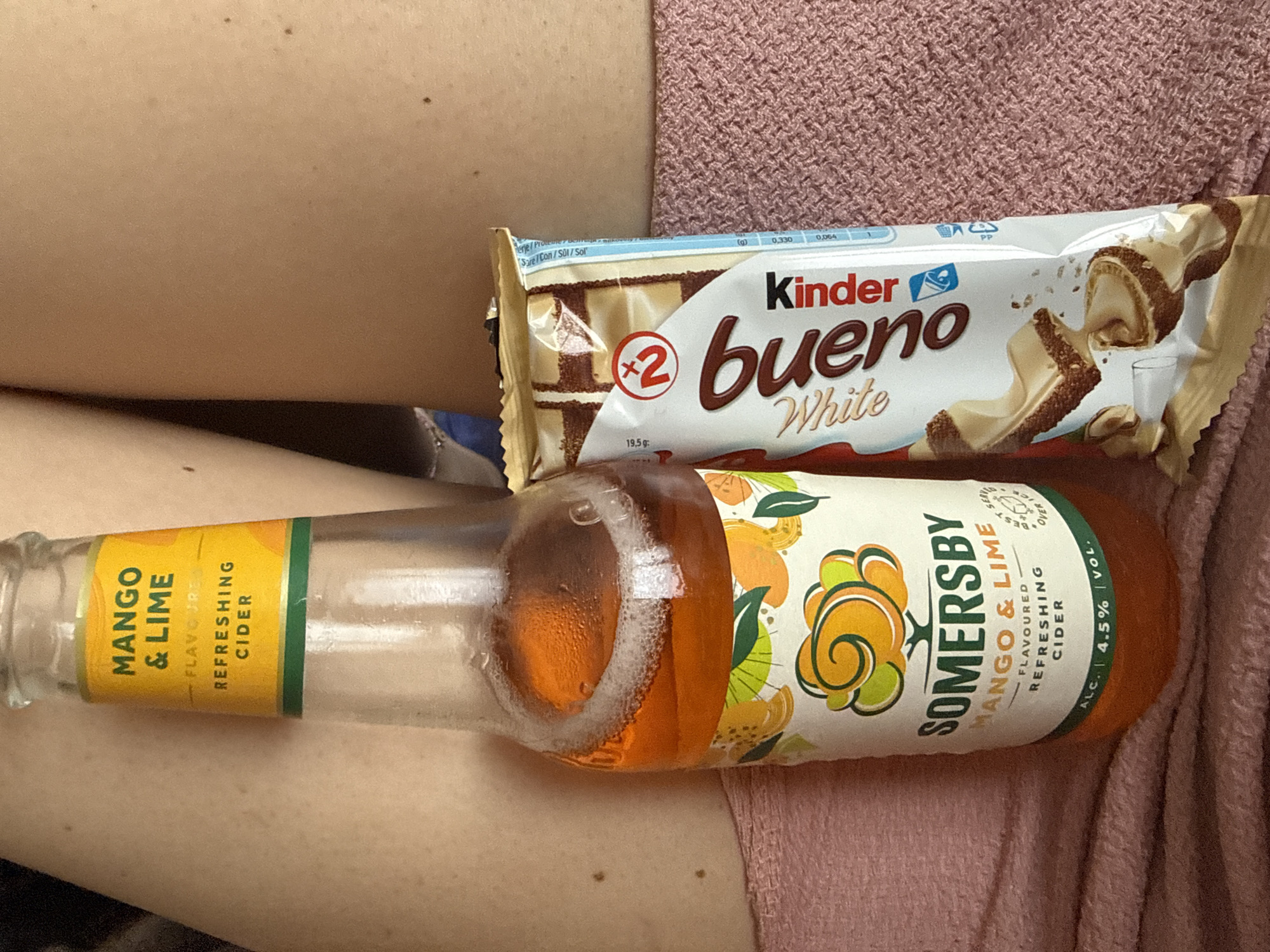 The image displays a close-up indoor scene featuring a bottle of flavored cider and a chocolate bar resting on a pink, textured fabric.
A clear glass bottle of Somersby Mango & Lime flavoured refreshing cider, containing an orange-hued liquid with some white foam at the top, is positioned centrally on the left. The label clearly indicates "ALC. 4.5% VOL." The bottle is partially consumed.
To the right of the cider bottle is a white and brown packet of Kinder Bueno White chocolate bars, featuring an image of broken chocolate pieces and the text "x2" and "19.5g".
The items are placed on a soft, possibly knitted or fleece, pink material, which appears to be a blanket or clothing. In the background, behind both objects, visible human skin, likely a thigh or upper arm, is present. The skin has several small, dark moles or freckles. A glimpse of a darker, possibly blue, item or fabric is partially visible in the upper left background.
No specific geographical identifiers are visible within the image to determine a location such as Kazanlak, Bulgaria.