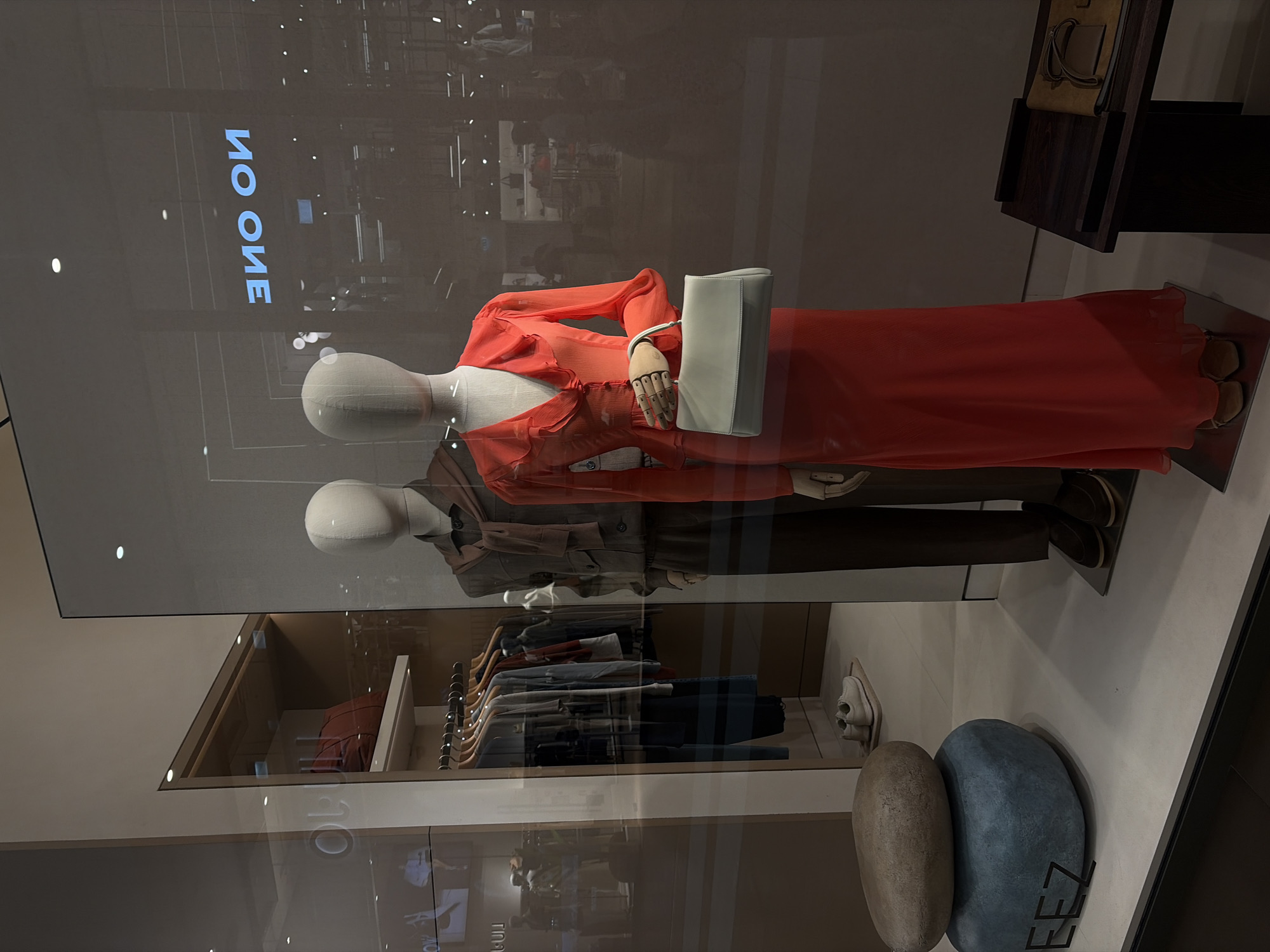 This is a full-shot, eye-level, medium-width photograph of a clothing store window display in Saint Petersburg, Russia. The display features two mannequins. The mannequin on the left is male, wearing a taupe suit jacket, a brown scarf draped around its neck, and dark brown pants. The mannequin on the right is female, wearing a vibrant orange dress with ruffled sleeves and a V-neck. It holds a pale blue rectangular handbag. Behind the mannequins, clothes hang on a rack, and further back, shelves are visible with bags and other accessories. In the foreground, a display consists of two stacked decorative stones, the bottom one blue and the top one brown. A portion of the store's signage, "EEZ", is visible on the blue stone. Reflections in the glass show the interior of the store, including lights and other displays, as well as an illuminated sign reading "NO ONE" in blue, backward. The floor is a light-colored tile.