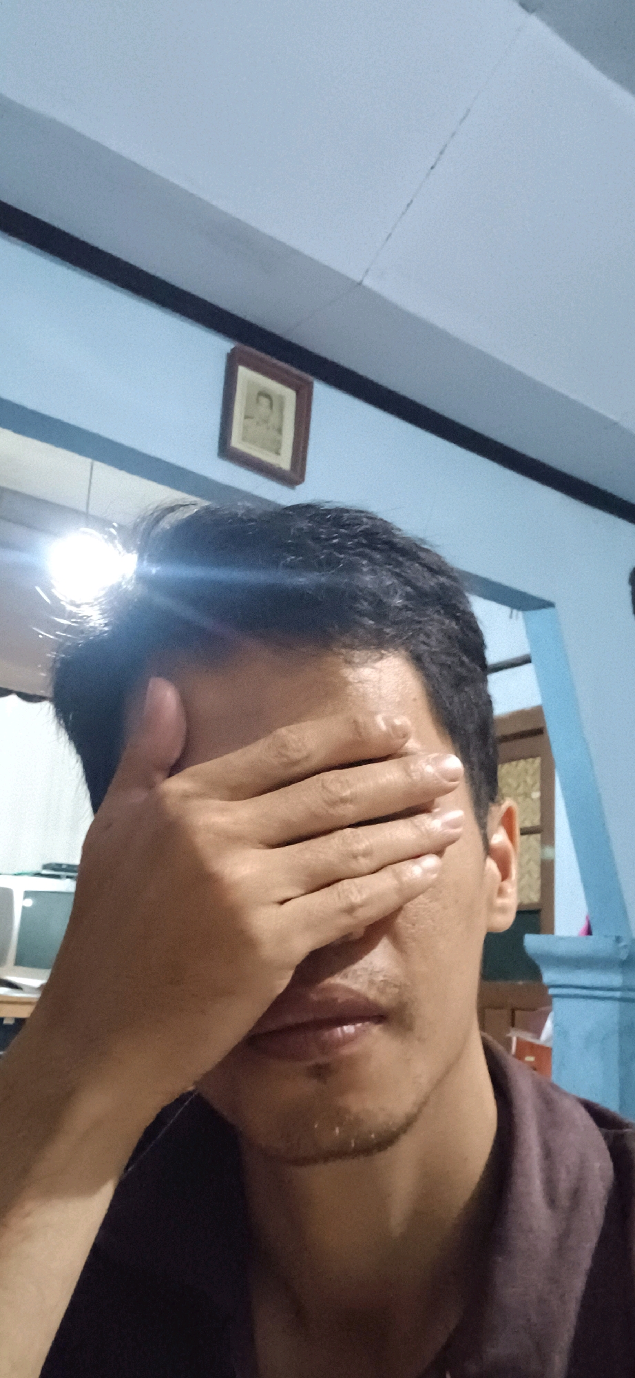 A medium shot captures a person covering their eyes with their right hand, obscuring the upper half of their face. The individual has dark hair and a mustache. They are wearing a dark-colored collared shirt. The background consists of a light blue wall with a dark horizontal trim near the ceiling. A framed picture hangs on the wall. To the left, a portion of a computer monitor is visible. The image appears to have been taken indoors, likely in a residential setting in Wonokerso, Indonesia.