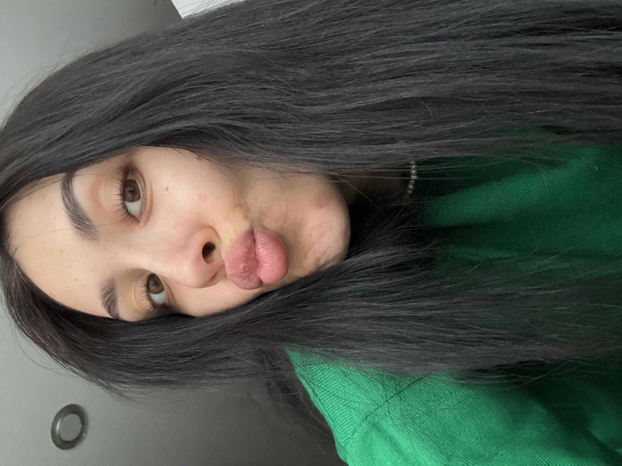 A young woman with dark hair and brown eyes is angled in the frame. She is wearing a green sweater, and making a kissing face. There is a light-colored wall visible in the background. The location is not discernible in this image. The image does not contain any information linking it to the city of Votkinsk, Russia.