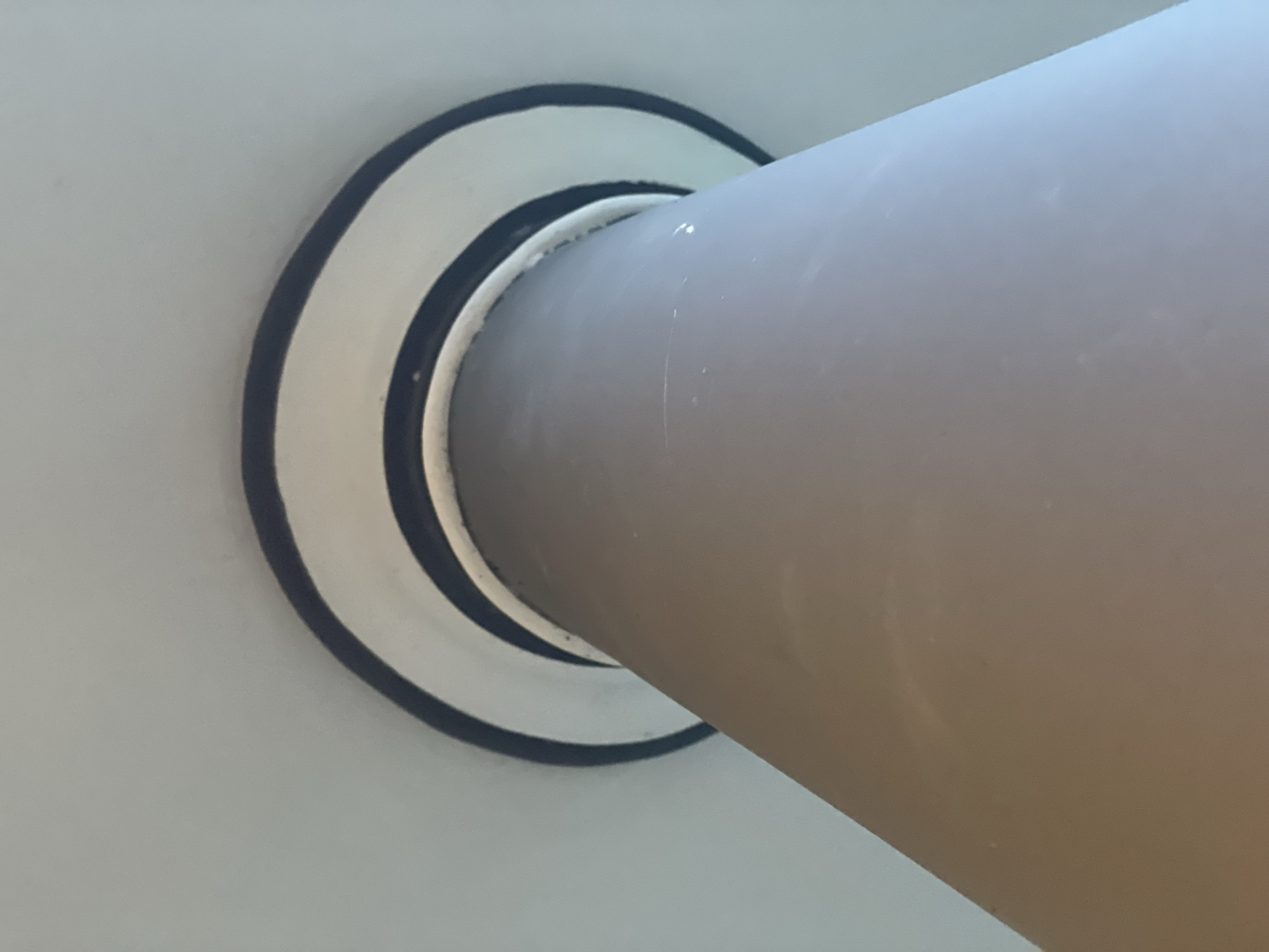 A low-angle, close-up shot looks up at a cylindrical, beige-colored pillar that tapers slightly as it rises. The pillar is positioned centrally in the frame. At the top of the pillar, where it meets the ceiling, there are two concentric rings, each with a dark blue or black outer edge and a lighter inner ring, likely painted. The ceiling is a light, neutral color, possibly white or off-white, and appears to curve gently around the pillar. The lighting is soft and diffused, casting minimal shadows. The overall impression is one of architectural detail, possibly within a building or structure. No people or discernible actions are present in the image. The specific location in Phan Thiet Ward, Vietnam, is not indicated by any visual cues in the photograph.