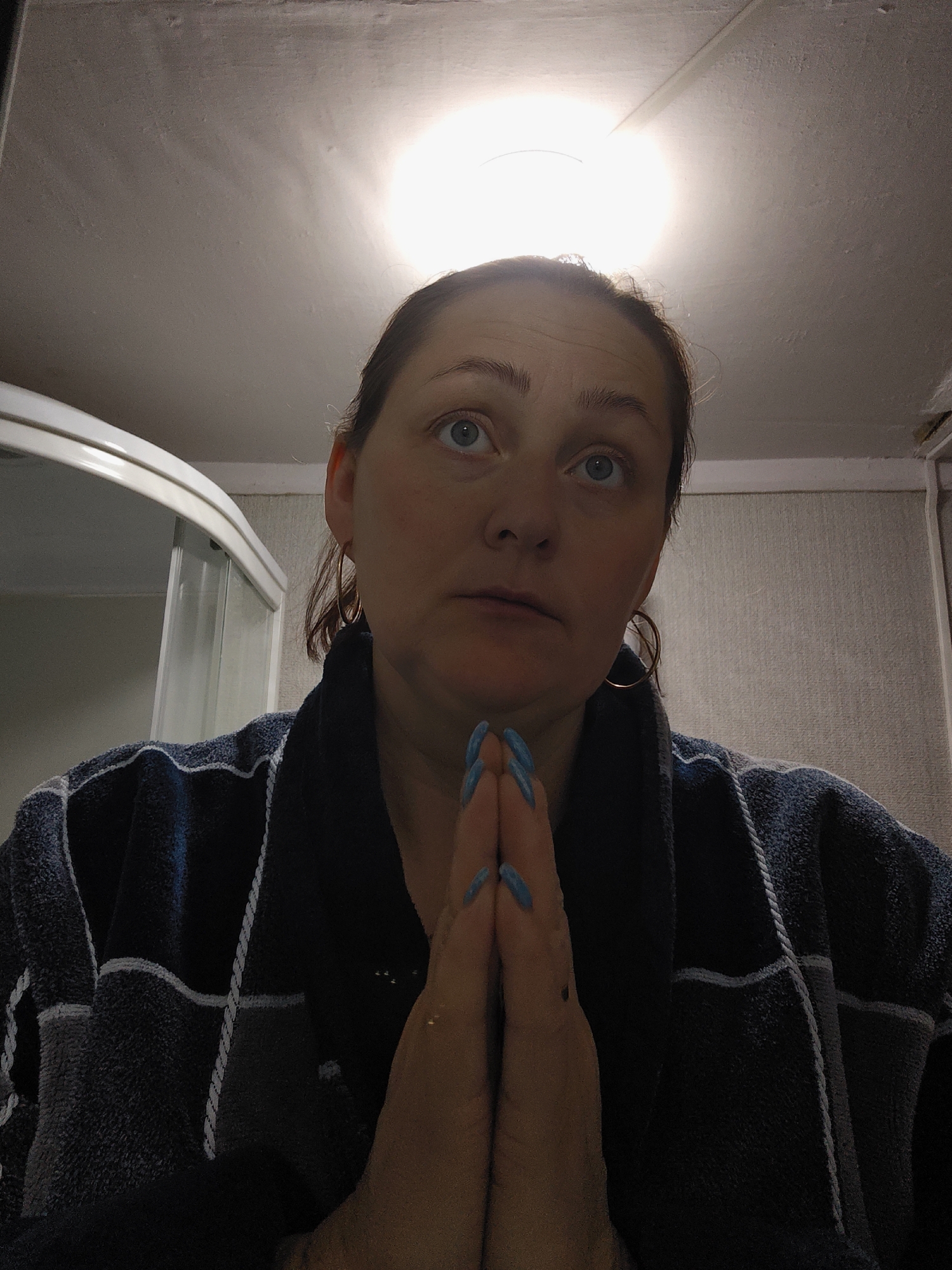 A head-and-shoulders portrait captures a woman indoors, presumably in a bathroom in Фоки, Russia. She is looking upwards and slightly to her right, directly towards a bright, circular light fixture on the ceiling that appears to be the primary light source in the room. Her hands are clasped together in a gesture commonly associated with prayer or earnest thought, positioned in front of her chest. Her expression is serious and contemplative, with her light-colored eyes (blue/grey) wide and focused.
The woman has brown hair, which appears to be pulled back, and she is wearing large, thin gold hoop earrings. She is clad in a dark blue or black bathrobe with distinct lighter blue or grey striped patterns. Her fingernails are painted a vibrant bright blue.
The background shows a plain, light-colored ceiling with some texture and what looks like a seam or crack. To the left, a portion of a white, curved structure, likely part of a shower cabin or bathtub enclosure, is visible. The wall behind her on the right is also light-colored and textured, possibly with wallpaper or an uneven finish. The artificial light casts a warm glow, illuminating her face from above. There is no visible text in the image. The time of day is not discernible due to the indoor artificial lighting.