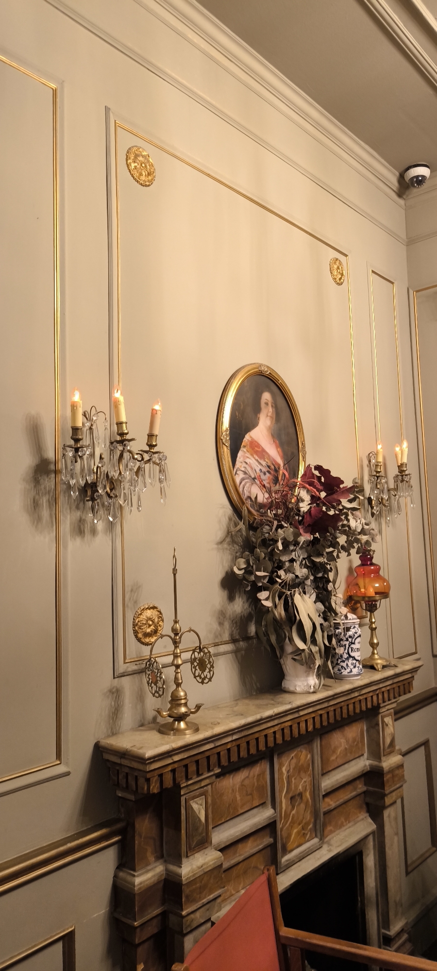 An interior scene depicts a room featuring ornate wall decor, including panels with gold trim and medallions. A marble-topped mantel supports an arrangement consisting of a portrait, candelabras, a brass oil lamp, a vase with flowers, and a decorative blue and white container. A red chair is partially visible in the foreground. A surveillance camera is present in the upper right corner. There is no indication of a location in Barcelona, Spain in this image.