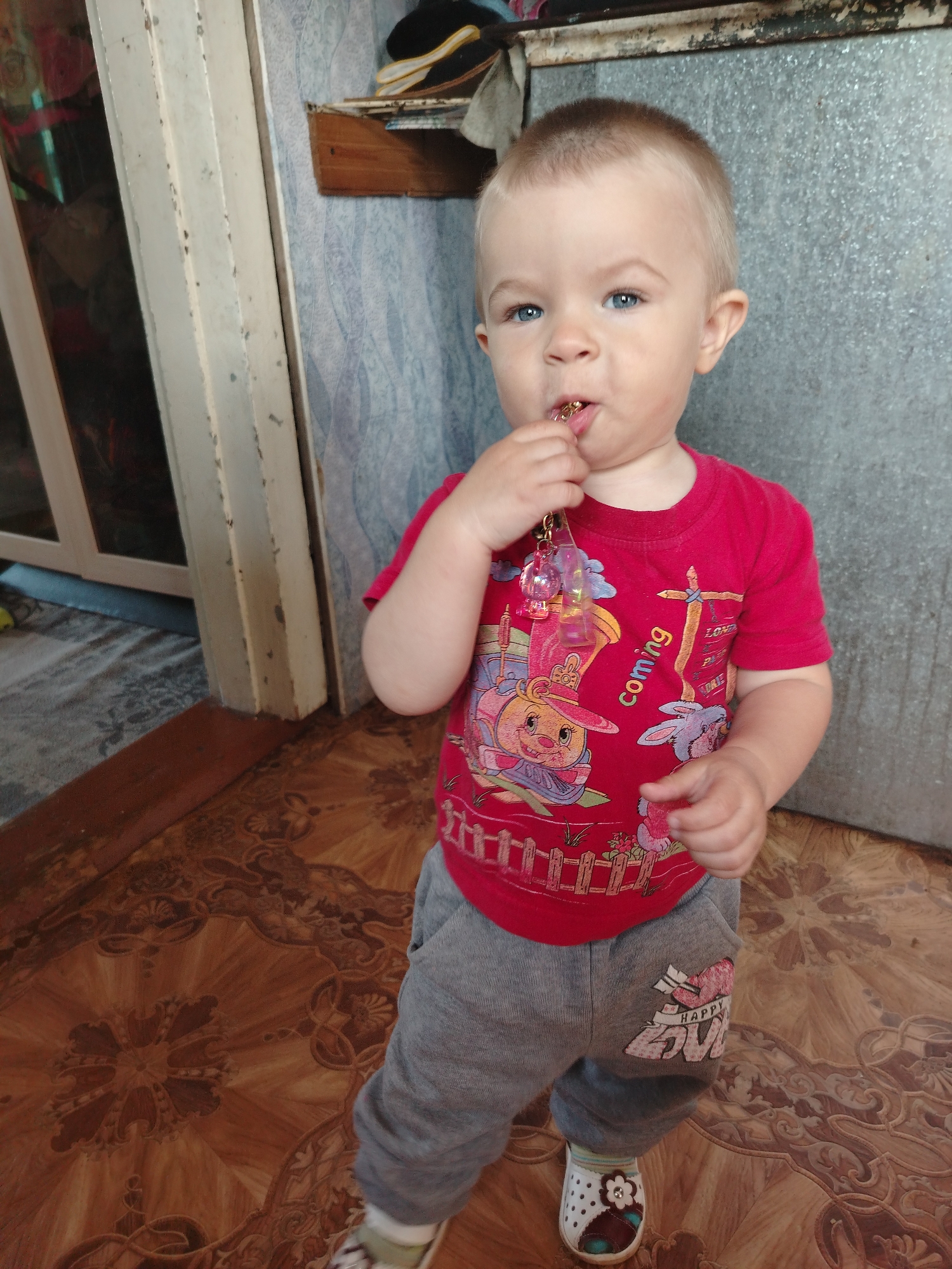 A light-skinned toddler with blonde hair and blue eyes is standing on a patterned brown linoleum floor, facing the camera and chewing on a keychain. The toddler wears a red t-shirt with colorful cartoon characters and text that reads "coming" and "HAPPY LOVE". Gray sweatpants are worn with white and brown shoes adorned with a flower. In the background, a metal wall and a door frame are visible, along with a shelf holding a black and yellow hat. This scene is likely in a residential interior in Zheshart, Russia, given the linoleum flooring and common household elements.