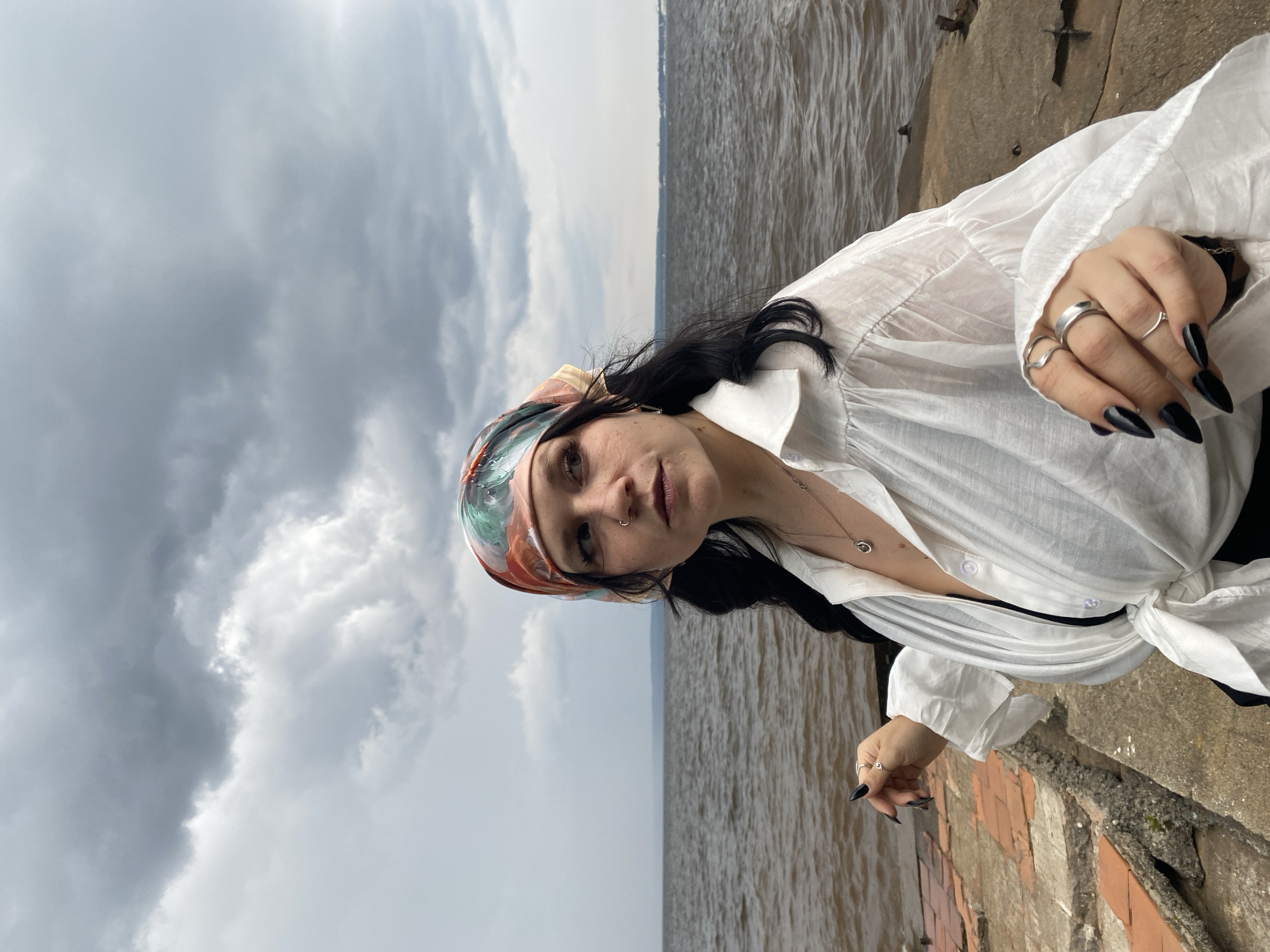 A woman with dark hair and a colorful headscarf stands by the Kama River in Chaikovsky, Russia. She is wearing a white open-collared shirt tied at the waist, exposing a black undergarment. The sky is overcast with dark clouds, and the river water is choppy. She is adorned with rings on her fingers and a necklace. The foreground shows a concrete and brick embankment.