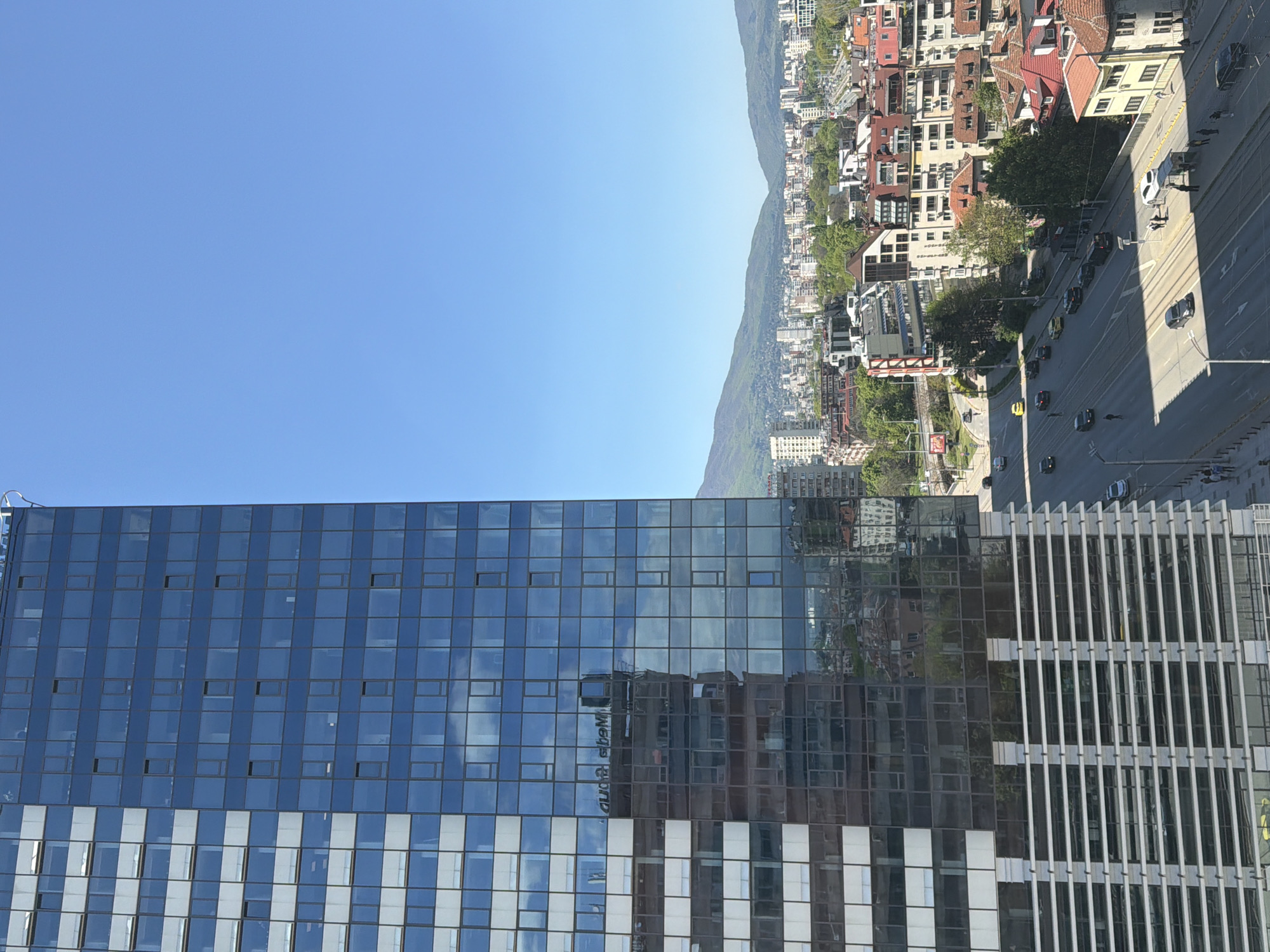 The image captures a view in Sofia, Bulgaria. A modern glass-clad building with horizontal white stripes on the lower part dominates the left side of the frame. The building reflects the urban landscape and sky, including the letters 'A.M.S.' reversed.
Beyond the building, there's a cityscape of Sofia, featuring various buildings with tiled roofs, interspersed with trees. A mountain range provides a scenic backdrop. A major thoroughfare cuts through the city with cars visible. Some pedestrians walk on the sidewalks. The sky is clear.
