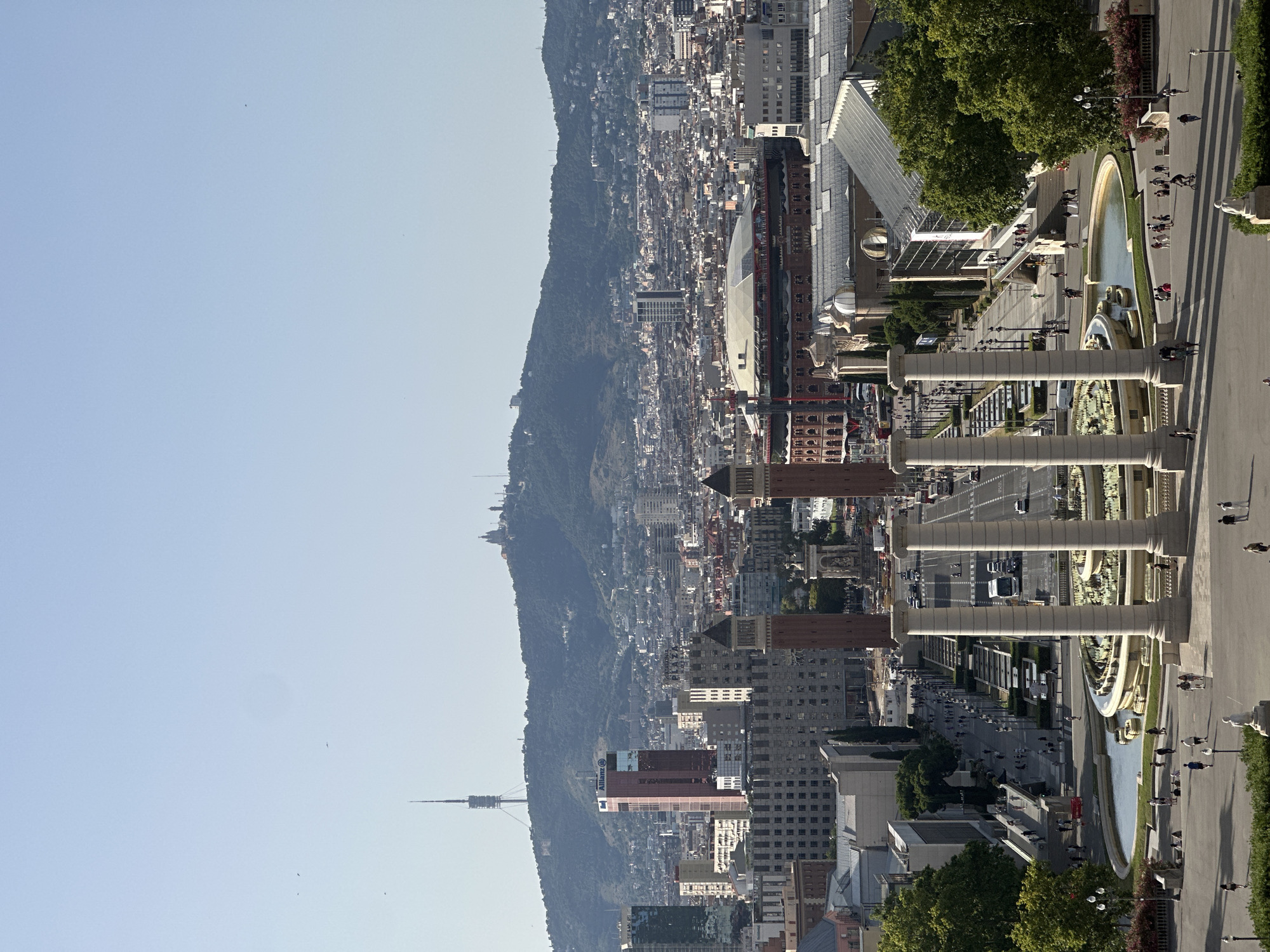 This is an aerial view of Barcelona, Spain, focusing on the Plaça d'Espanya and the Montjuïc hill in the background. In the foreground, four large classical columns stand prominently, framing a circular fountain with tiered water features. A wide, tree-lined avenue stretches away from the fountain, with pedestrians walking along the sidewalks and several cars and buses moving on the road. Further down the avenue, two large, ornate towers with arched windows are visible, likely part of the Venetian Towers. Beyond these towers, a sprawling cityscape unfolds, characterized by a dense collection of buildings of varying heights and architectural styles. In the middle ground, a large, domed structure, possibly an arena or exhibition hall, is discernible. Dominating the background is the lush, green Montjuïc hill, topped with several structures, including a prominent communication tower on its western slope and a chapel or religious complex on its eastern summit. The sky is clear and a pale blue, indicating a sunny day. The overall impression is one of urban grandeur and a blend of classical and modern architecture set against a natural landscape.