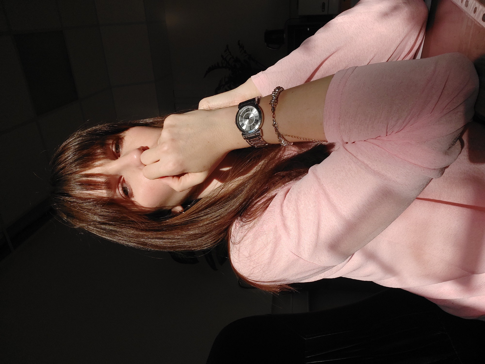The image presents a close-up, angled portrait of a person, likely female, with long, straight dark brown hair and light-colored eyes. She is wearing a light pink long-sleeved top. Her head is tilted, and her left hand is raised to her face, partially obscuring her nose and mouth, while her eyes look towards the viewer. On her right wrist, a silver-toned wristwatch with Roman numerals (such as IV and X) on its dial is visible, along with a matching silver-toned bracelet.
The scene is indoors, characterized by high-contrast lighting. Strong, direct sunlight enters from the right, illuminating parts of her hair, arm, and the pink fabric, casting sharp, deep shadows across the rest of the subject and the background. This creates a dramatic effect, with most of the background appearing as a dark, unidentifiable space, though subtle patterns suggesting a tiled ceiling are faintly visible in the upper left. The strong, directional light suggests it is daytime, likely morning or late afternoon. The person's expression is somewhat obscured by her hand. The location context provided is Chaikovsky, Russia. There is no clearly legible text other than the watch numerals.