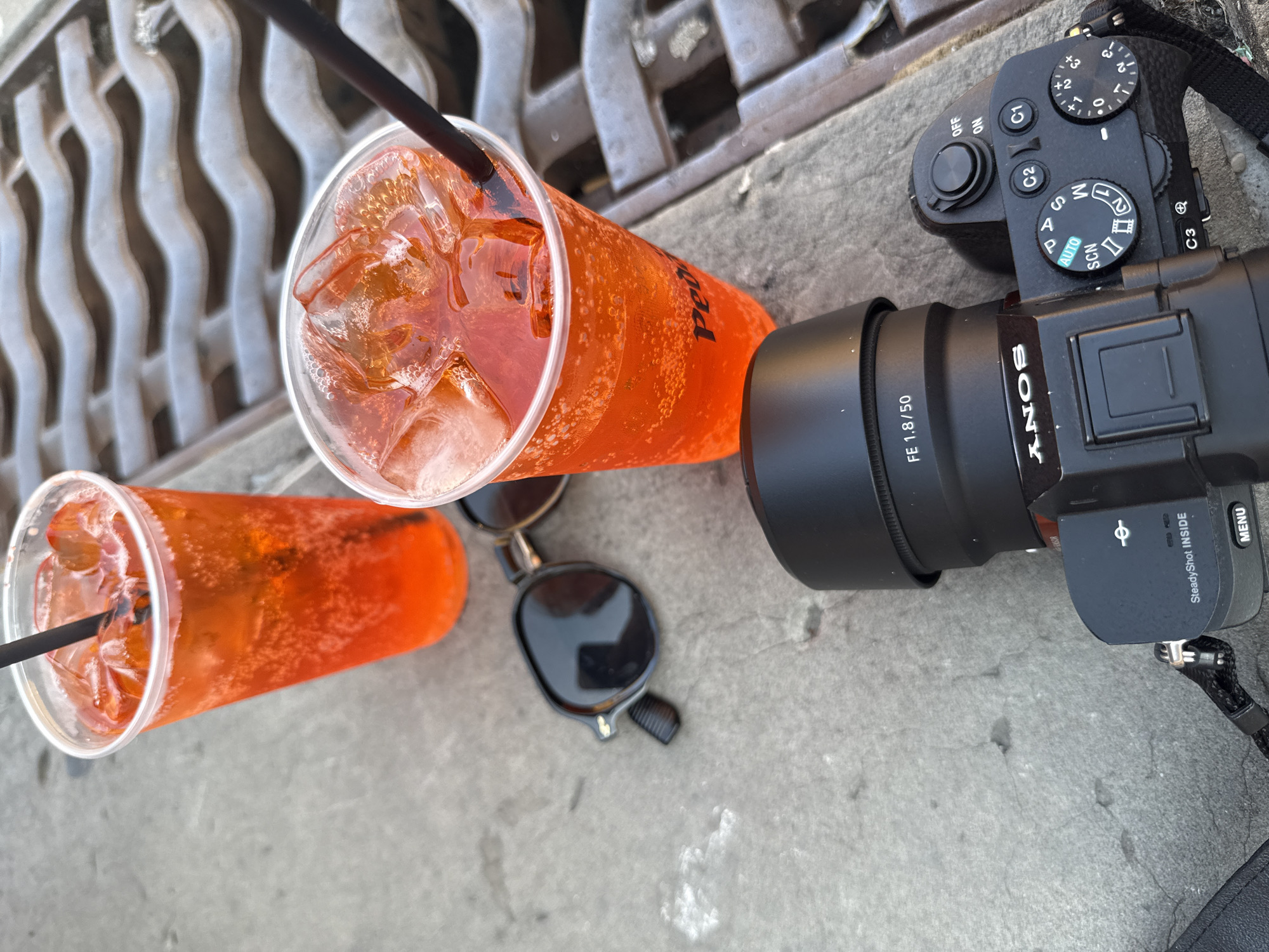 Two plastic cups of orange beverages with ice and black straws sit on a concrete surface next to a metal grate, likely a storm drain. A pair of black sunglasses rests between the cups and a black Sony camera with a lens is positioned in the foreground. The camera has text that reads "FE 1.8/50" on the lens and "SONY" and "SteadyShot INSIDE" on the body. The dial on top of the camera shows various shooting modes, including "AUTO," "SCN," and "M." The overall scene suggests a casual outing in an urban environment, possibly during warm weather. The presence of the camera implies someone is documenting their experience. The metal grate is characteristic of Florentine street infrastructure.