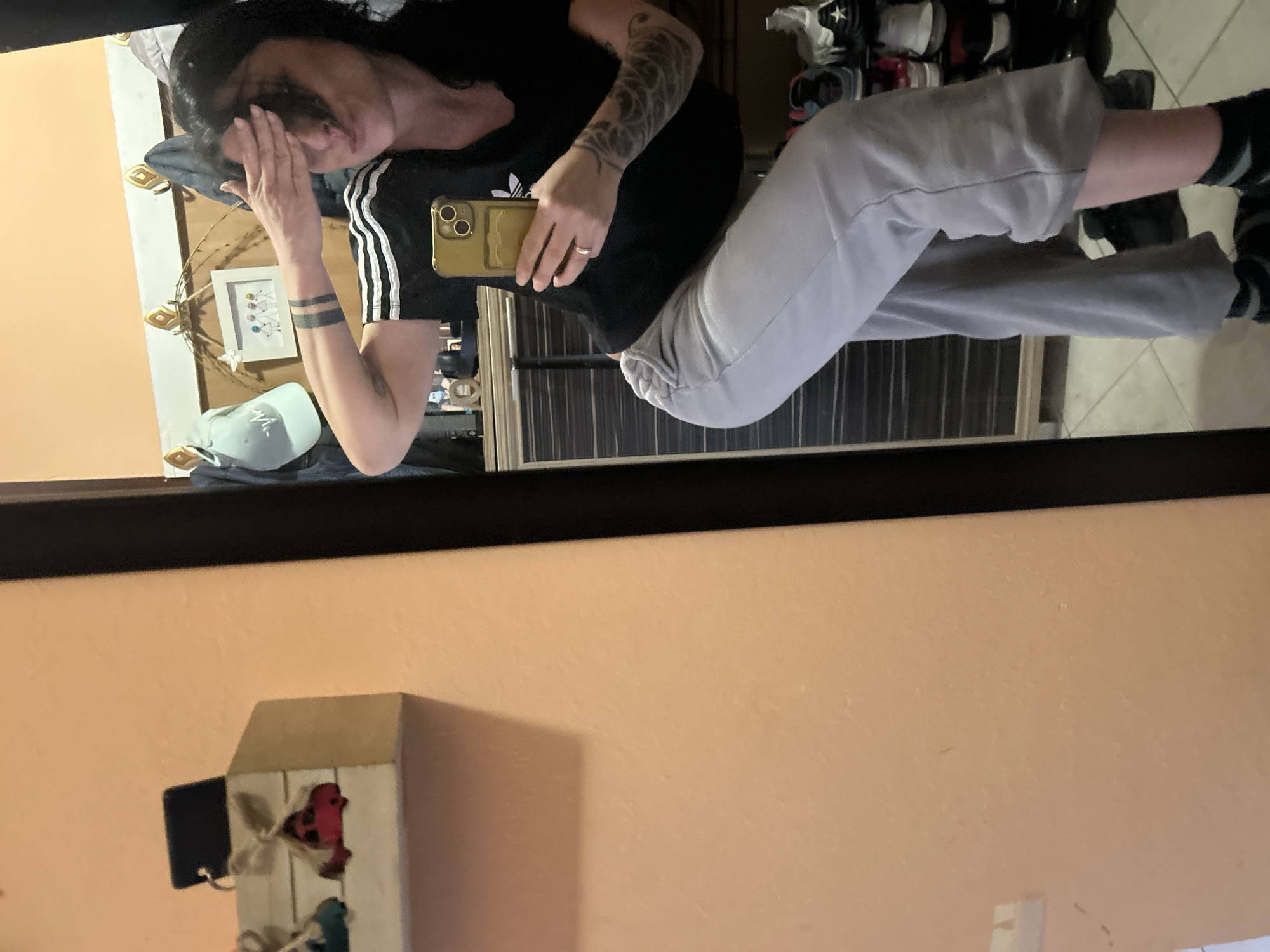 The image appears to be a reflection of a woman taking a selfie in front of a mirror. She is wearing a black t-shirt with white stripes on the sleeves and grey sweatpants. She is holding a gold-colored smartphone in her right hand. Her hair is dark and she has tattoos on her left arm. In the background, there is a shoe rack with multiple pairs of shoes, a piece of furniture with dark brown and light brown paneling, a baseball cap, and a framed picture. The reflection also shows a light peach-colored wall with a decorative birdhouse hanging on it. The city and location are unknown.