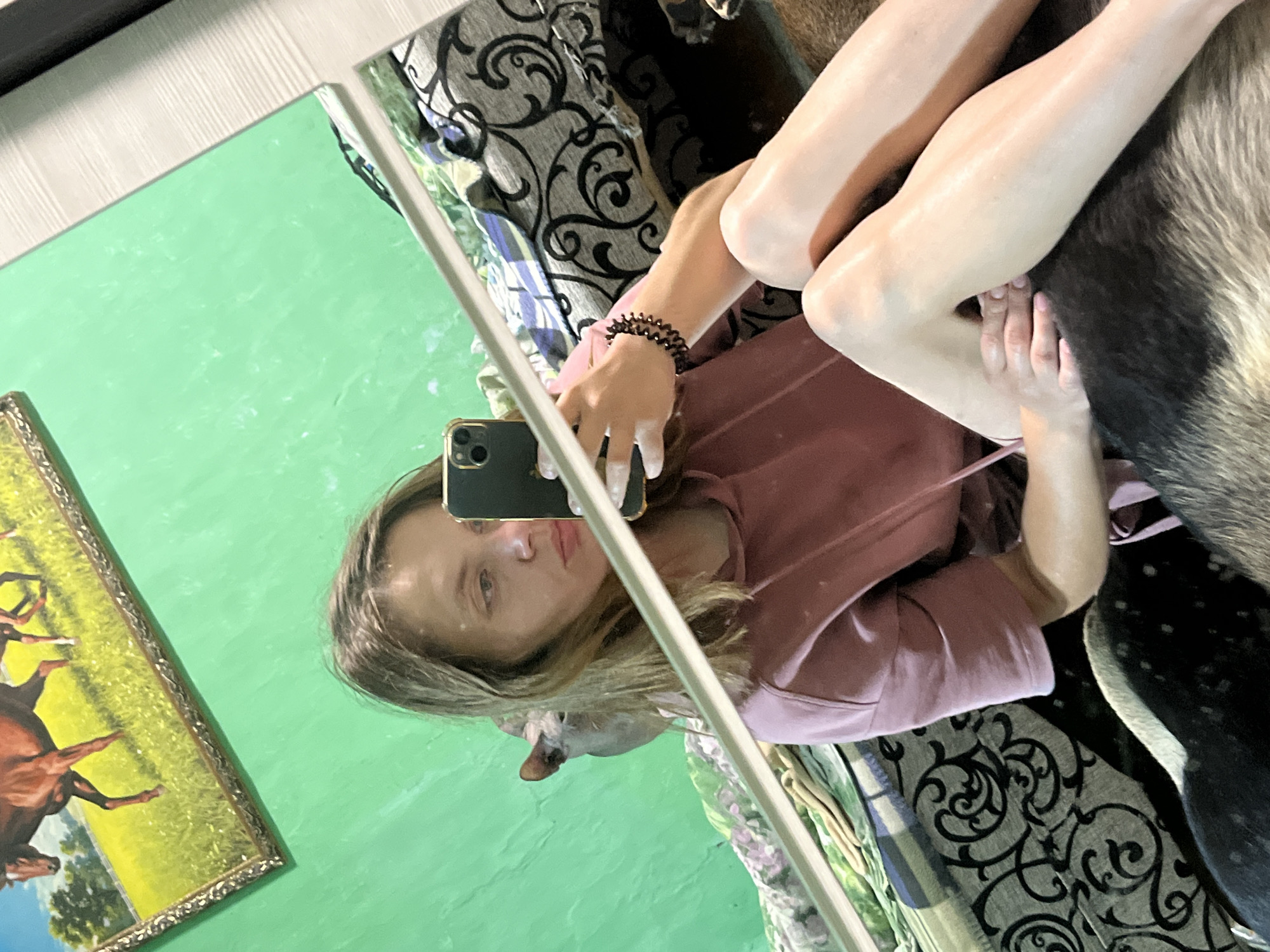 A young woman with shoulder-length blonde hair is taking a mirror selfie in what appears to be a bedroom. She is wearing a pink hooded sweatshirt and is seated on a bed with a patterned comforter. Her left arm is resting on her leg, and she is holding her phone with her right hand. In the background, a framed painting of horses is visible on a bright green wall. A cat is partially visible behind the woman's head in the reflection. The overall impression is one of a casual, domestic setting. There are no specific indicators that this scene is located in Nizhny Tagil, Russia.