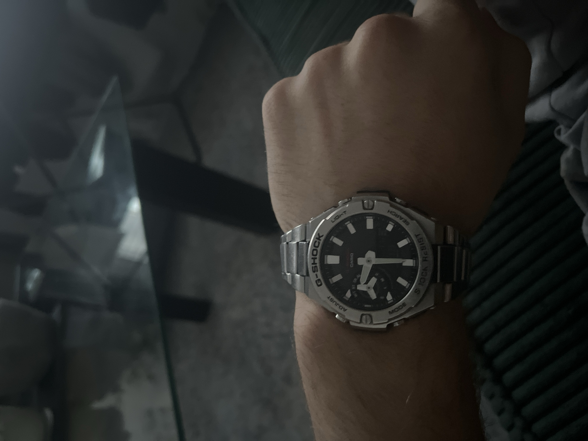 This media file captures a close-up shot of a person's left wrist, adorned with a silver G-Shock watch. The watch features a black dial with white hour markers and hands, and a smaller dial at the bottom. Visible text on the watch includes "G-SHOCK", "TOUGH", "WATER RESIST 20 BAR", "LIGHT", "ADJUST", and "MODE". The background is blurred, showing a dark, textured surface, possibly a couch or chair, and a reflective surface, hinting at a glass table. The overall lighting suggests it might be evening or indoors. The scene appears to be a casual, everyday moment, with no specific event taking place. The context of Pruszcz Gdański, Poland is provided but not directly inferable from the image itself.