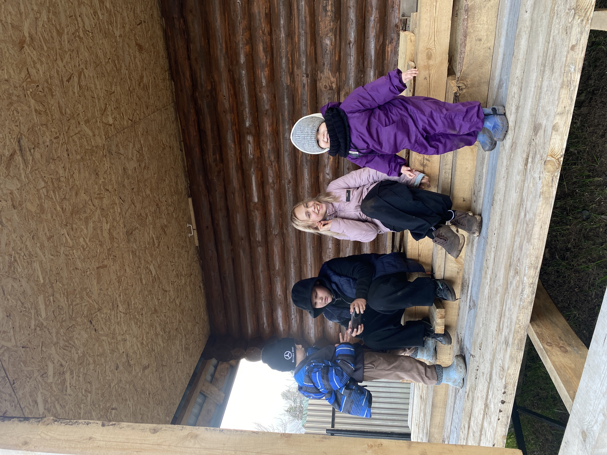 Four children are on wooden planks inside an unfinished wooden structure. The structure is constructed of timber and plywood. The child at the top wears a purple snowsuit and a grey hat. The next child down wears a light pink jacket and black pants. The third child wears a black coat and pants. The child at the bottom wears a blue jacket and tan pants. The location is Chaikovsky, Russia.