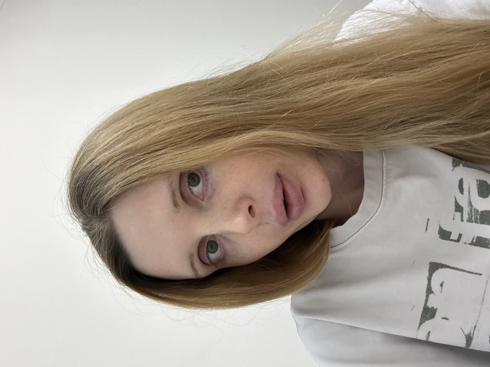 A young adult female with blonde hair and light skin is looking directly at the camera. Her eyes are blue and she has light makeup on her lips. She is wearing a light-colored t-shirt with a graphic print on the front. The background is a plain white wall. The image appears to be a selfie, taken at close range.