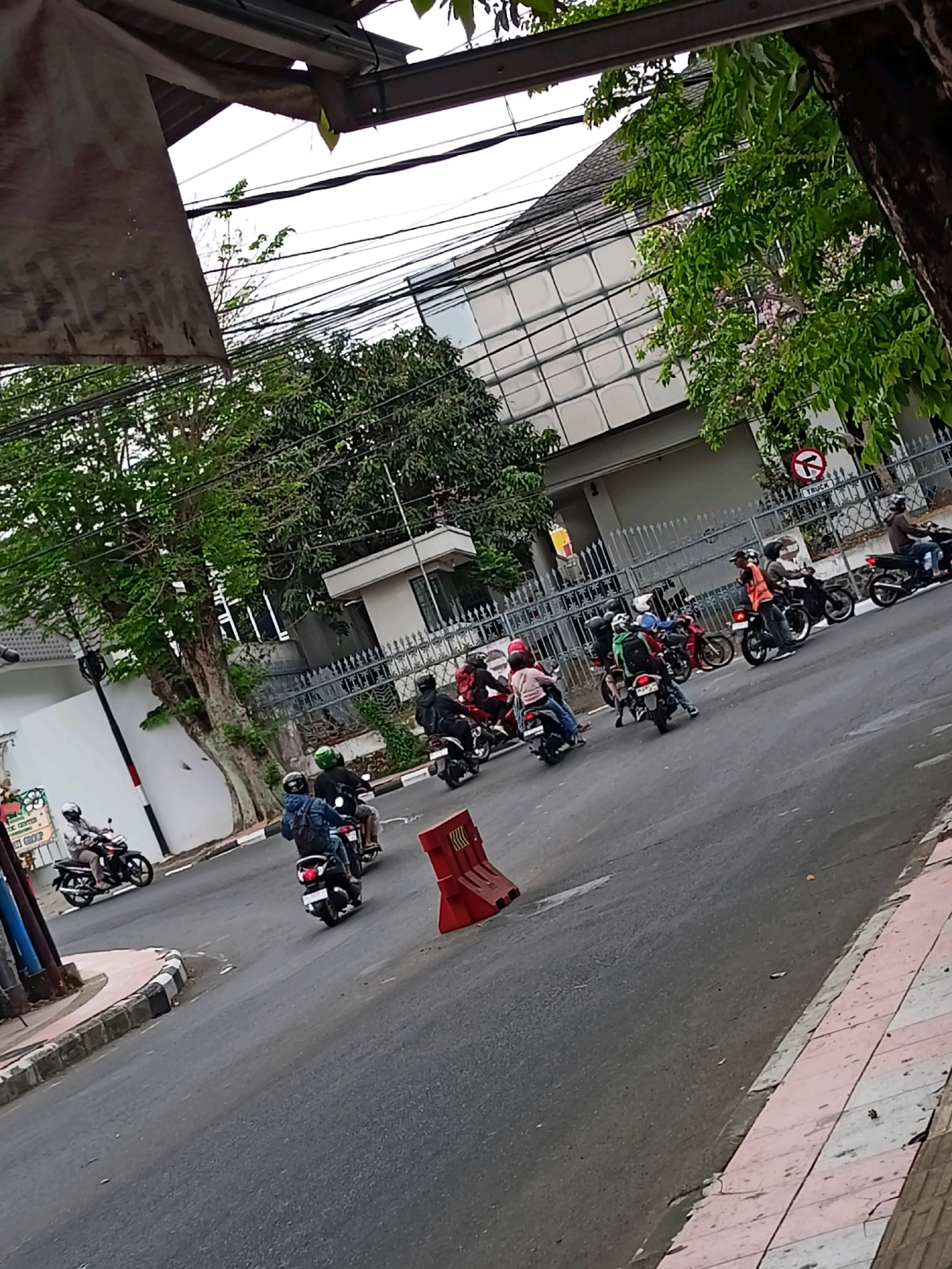 The image depicts a street scene in Magelang, Indonesia, with several motorcycles in motion. The road appears to be paved asphalt, with a sidewalk to the right featuring a decorative tile pattern in pink and white. On the left side of the road, there is a white building with a black and white striped pole in front of it. A partially visible sign with some text is also present on the left.
A group of motorcycles, each with at least one rider wearing a helmet, are traveling on the road. One motorcycle on the far left is heading towards the viewer, while others are moving away in the distance. A red traffic barrier is positioned on the right side of the road, near the center of the frame.
In the background, there are buildings and trees. A multi-story building with a checkered facade is visible behind a fence. Overhead, a network of power lines crisscrosses the sky. The overall impression is a typical urban street environment with active motorcycle traffic.