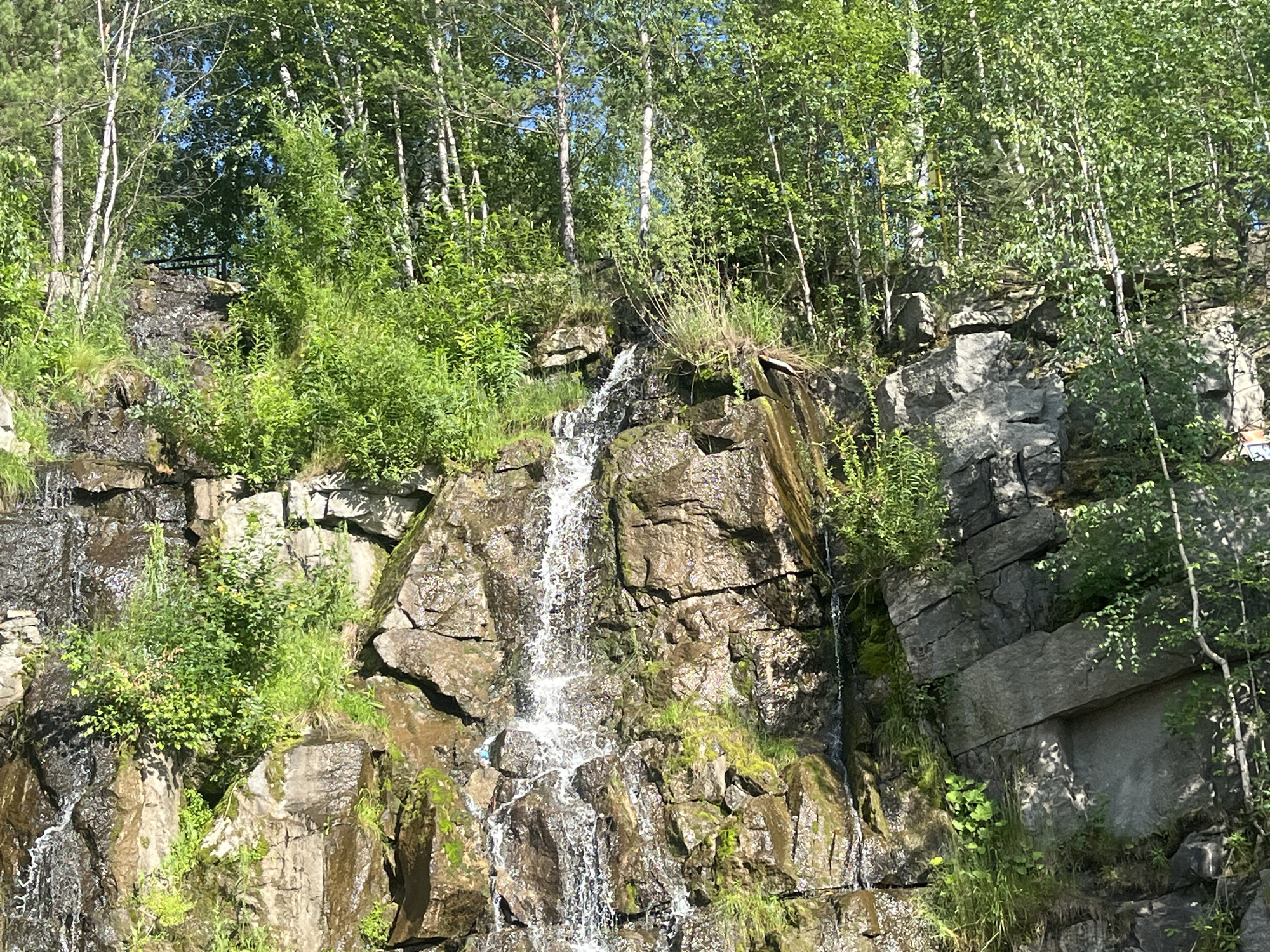 A narrow waterfall cascades down a rugged rock face surrounded by lush green foliage and tall trees. The water is a bright white against the dark, wet rocks, creating a dynamic contrast. Sunlight filters through the leaves, casting dappled shadows on the scene. Several deciduous trees with slender white trunks and full green canopies are visible at the top of the rock face, their branches reaching towards the sky. Patches of green moss and grass cling to the rocks, adding to the natural beauty of the environment. The rock formations themselves are varied, with some areas appearing rough and jagged, while others show more defined layers. The overall impression is one of a wild and serene natural landscape, likely captured on a clear day with pleasant weather. There is a railing visible at the top of the waterfall, suggesting this might be a natural attraction or park area. No people or text are visible in the image. The location context of Yekaterinburg, Russia, suggests this is a scene from the natural environment of that region.
