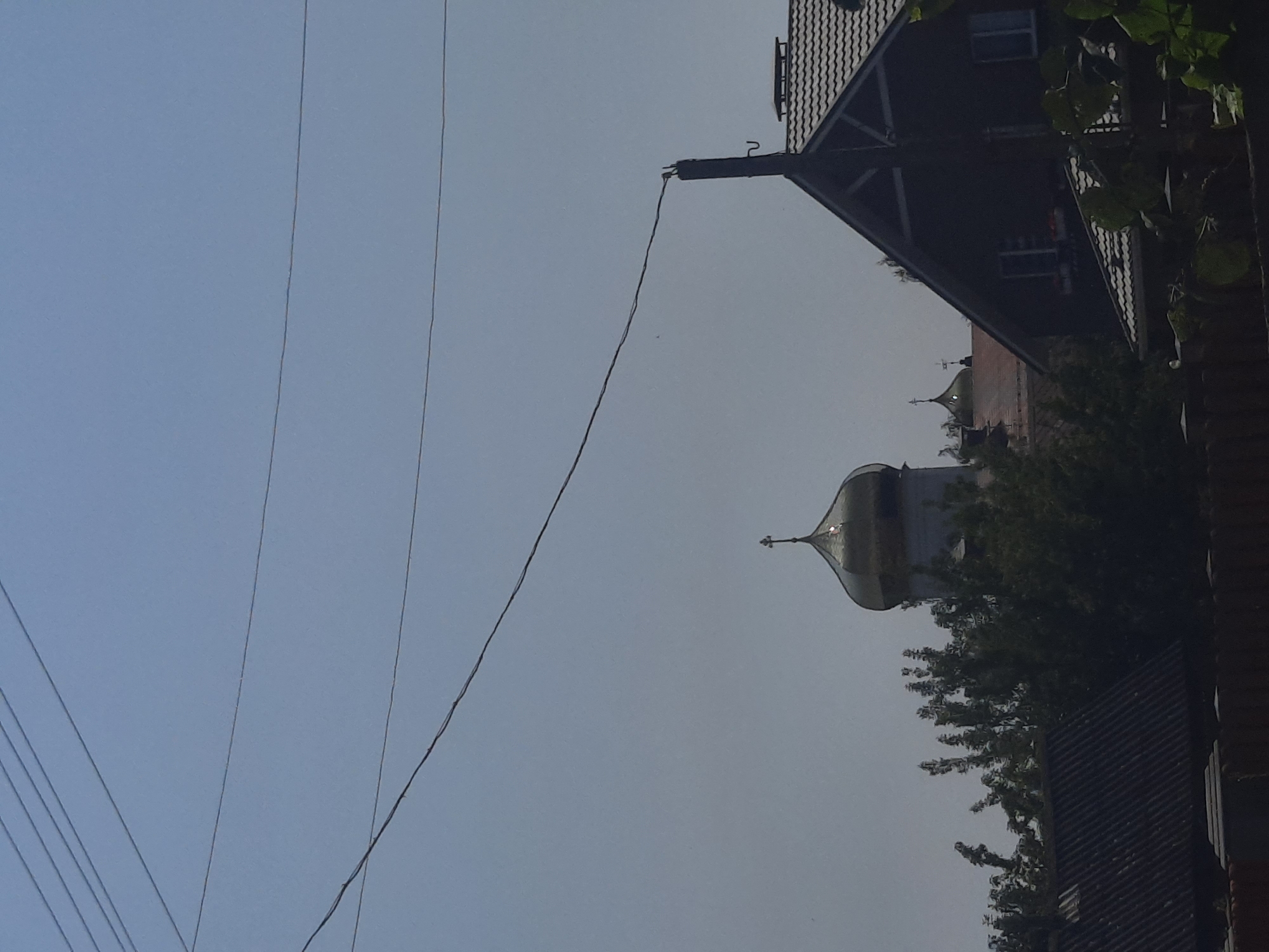 The image shows a bright, clear sky with multiple power lines stretching across the frame. In the middle ground, a large golden dome of an Orthodox church with a cross on top is visible, partially obscured by trees. To the right, a dark-colored house with a tiled roof and visible windows is present. The foreground features the top of a fence and some greenery. The scene suggests a residential area possibly on the outskirts of the city of Shklov, Belarus.