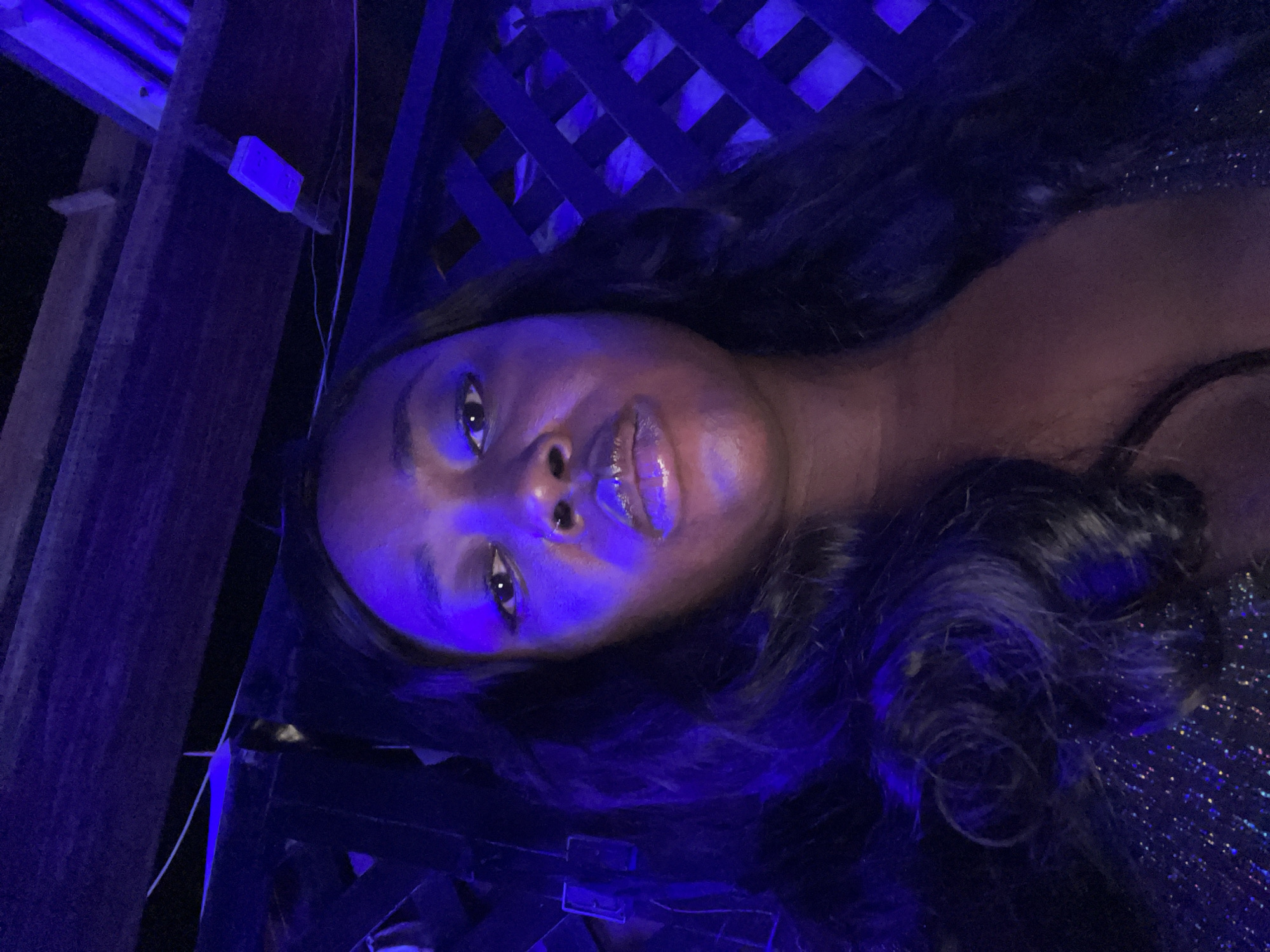 A single adult female subject is centered in a close-up, direct gaze selfie, illuminated by intense blue-purple ambient lighting, consistent with a nighttime entertainment venue. The scene is identified as being located in Abuja, Nigeria. The subject has dark skin, long dark wavy hair, and features glossy lips with a metallic sheen, and a small septum piercing. She wears a dark garment with visible glitter or sequins on the lower left. The background reveals dark wooden ceiling beams across the top of the frame and a dark decorative lattice structure on the right. Thin white wires and small rectangular fixtures are visible attached to the overhead structures in the low-light environment.
