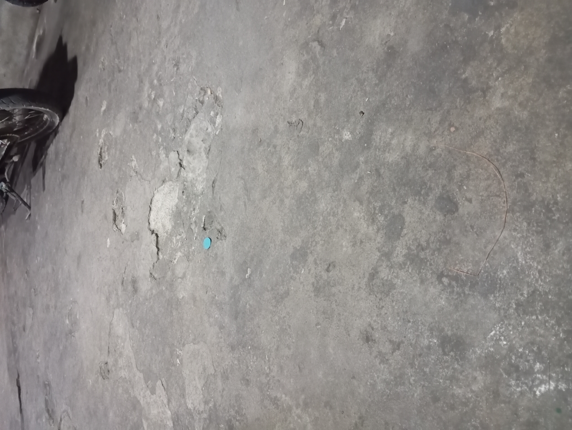 The image shows a section of a concrete floor in Rejosari, Indonesia, with a motorcycle parked in the background. The concrete surface is weathered and shows signs of damage, including cracks and chipped areas. A small, round, bright blue object is visible on the floor near the center-left of the image, appearing to be a cap or plug. In the lower portion of the image, a thin, curved, reddish-brown line is present on the concrete, which could be a piece of wire or string. The lighting suggests it might be evening or a dimly lit indoor space.