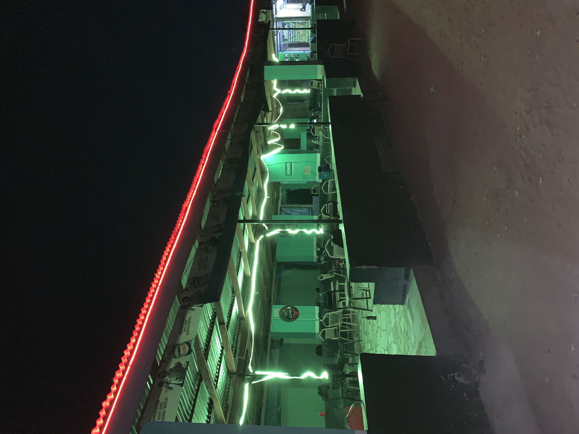 The image depicts an outdoor bar or eatery in New Stadium, Nigeria, at night. A long, low-rise structure with a corrugated metal roof, rimmed with red LED lights, houses the establishment. Fluorescent green string lights zigzag across the open-air seating area. Tables and chairs are arranged in rows, though few patrons are visible. The ground outside is dirt. Posters or advertisements are affixed to the building's facade. The sky is dark.