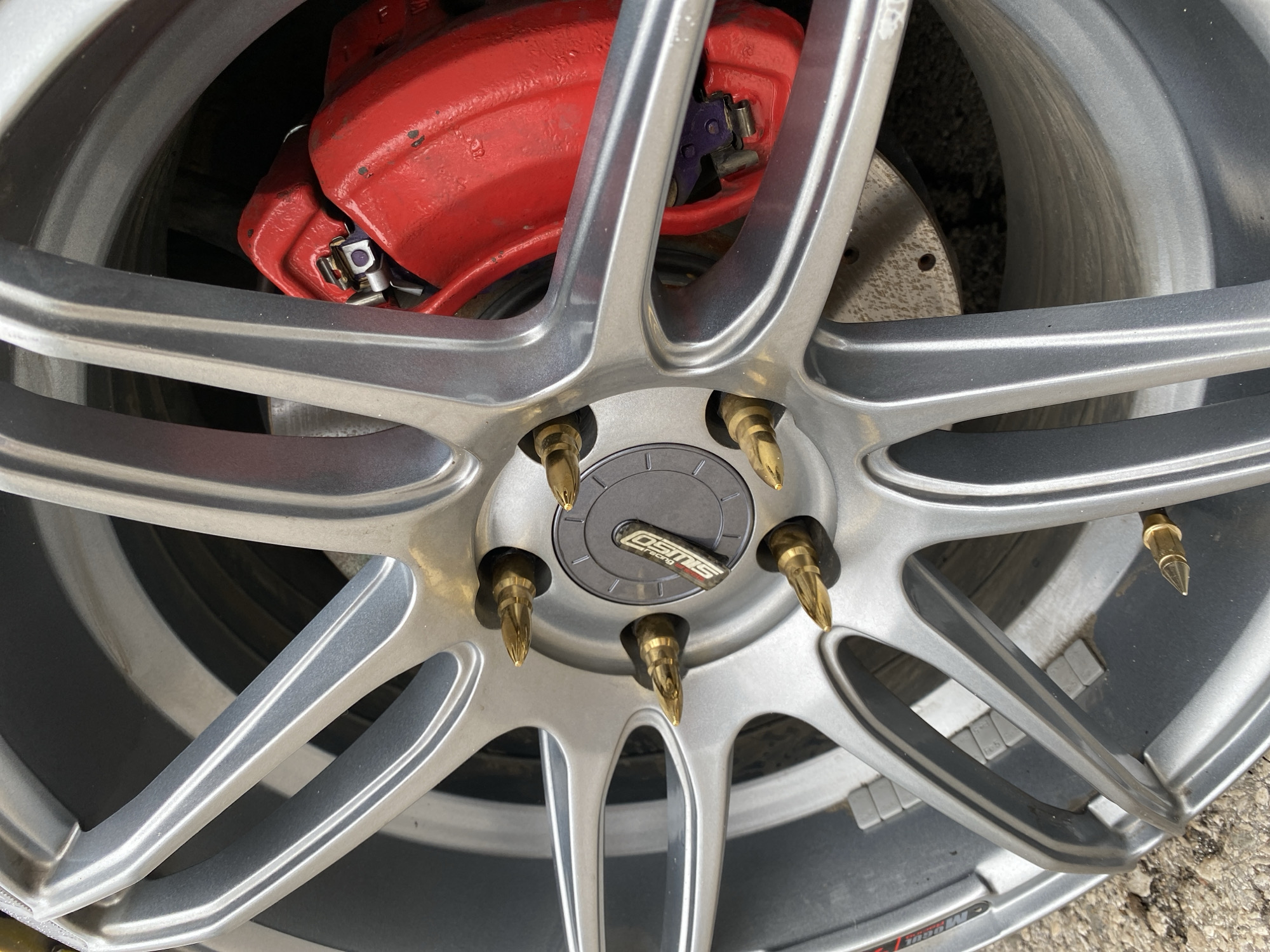 A close-up, eye-level, outdoor, horizontal shot showcases a silver custom car wheel with six pointed gold lug nuts. A bright red brake caliper is visible behind the wheel spokes. The background is dark, with the ground appearing to be gravelly and rocky, suggesting an outdoor setting, possibly a street or a repair shop. No people or animals are present. The image does not contain any information that would allow for its location to be specified in the city of Perm, Russia.