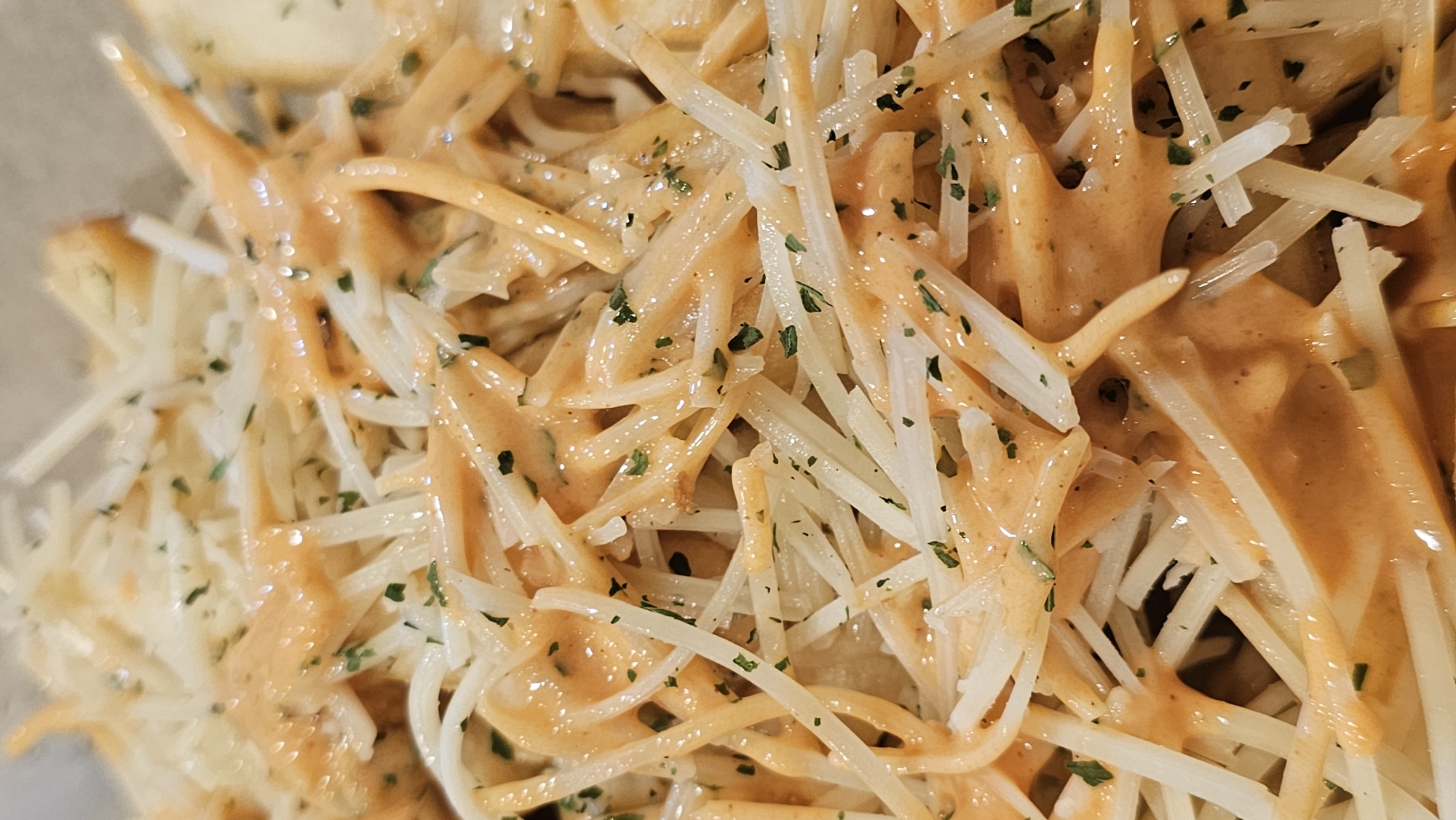 This is a close-up shot of a plate of food. The main components are long, thin, white strands resembling pasta or shredded cheese, covered in a creamy, orange-colored sauce. Green herbs are sprinkled over the dish. There are no people present. The location is unspecified, but it does not include the city of Puebla, Mexico.