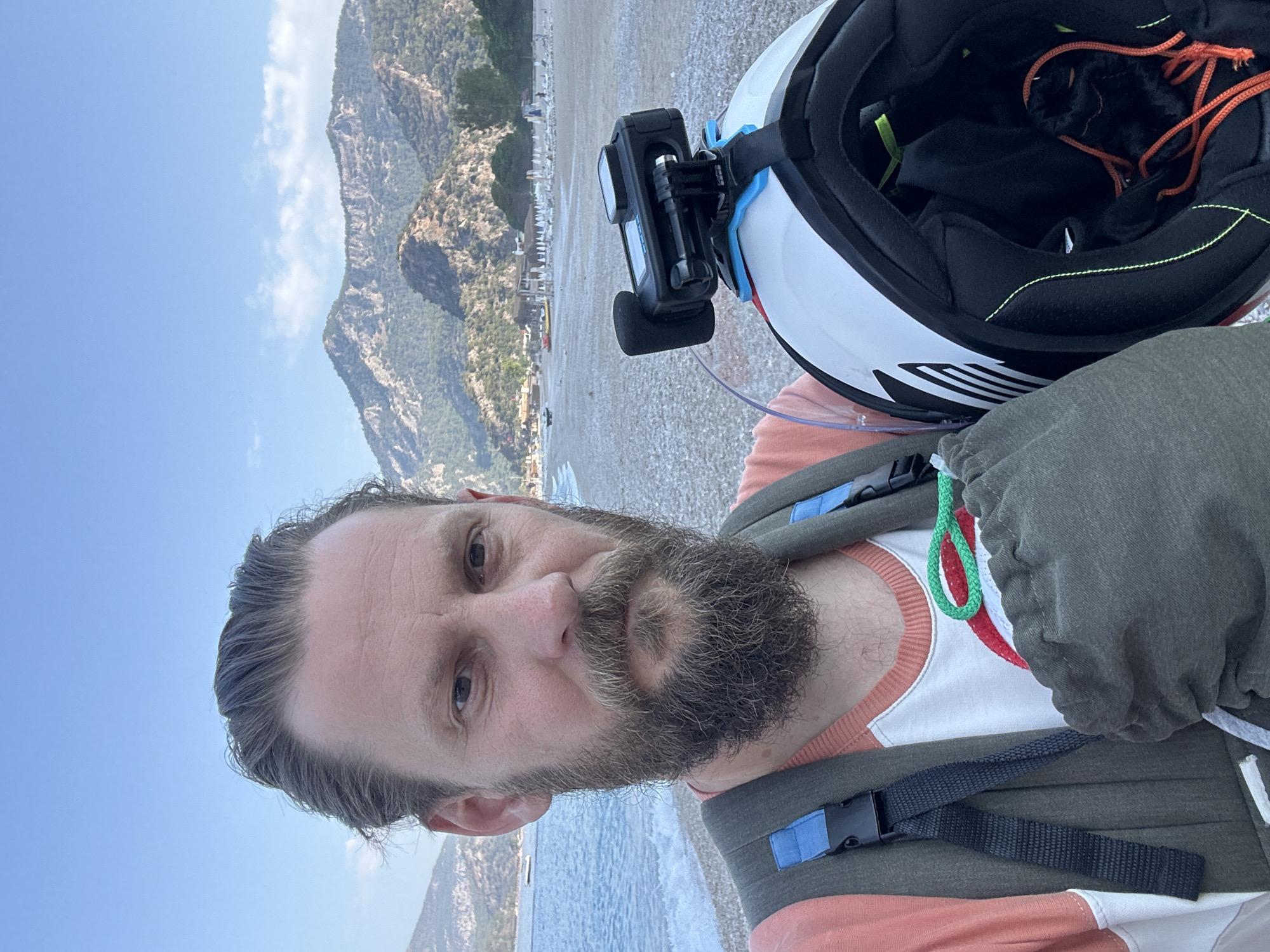 A man with a beard and a backpack is taking a selfie on a pebble beach in Fethiye, Turkey. He is holding a white motorcycle helmet with a GoPro attached to it. In the background, there is a body of water with a few boats, a beach with umbrellas and buildings, and mountains. The sky is blue with a few clouds. The man is wearing an orange and white t-shirt and a green vest.
