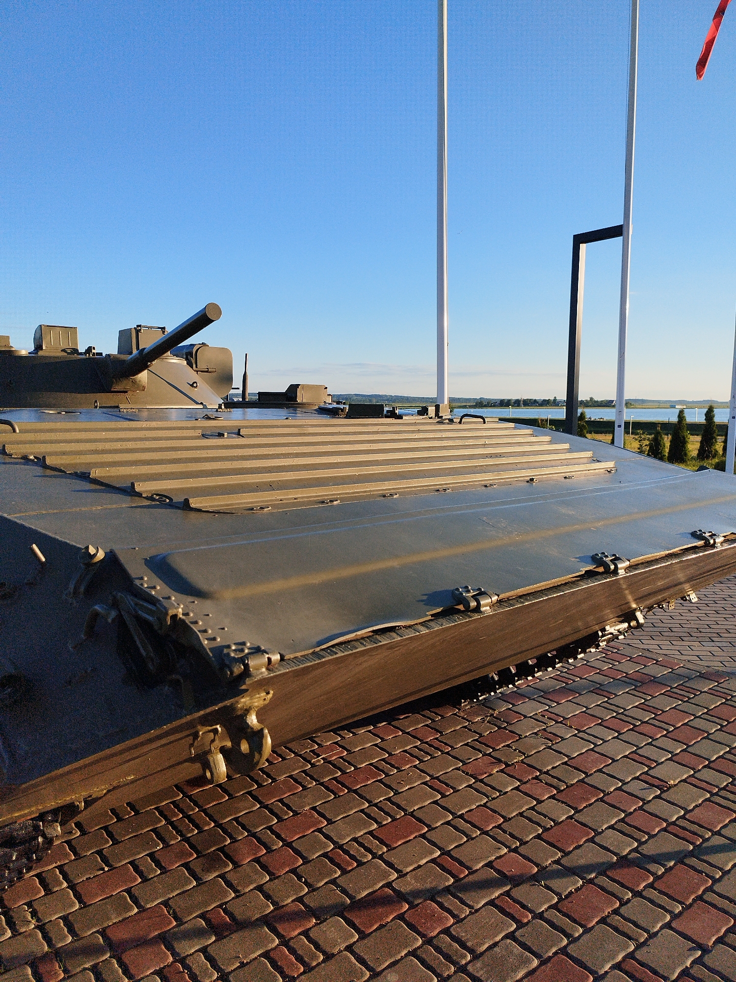 A military armored personnel carrier is parked on a paved walkway in front of a body of water under a clear blue sky. The APC is a military green color and has a gun turret. The walkway is made of interlocking pavers in shades of red and brown. In the background, there are several trees and some buildings. There are also flagpoles visible. The lighting suggests it is either early morning or late afternoon.