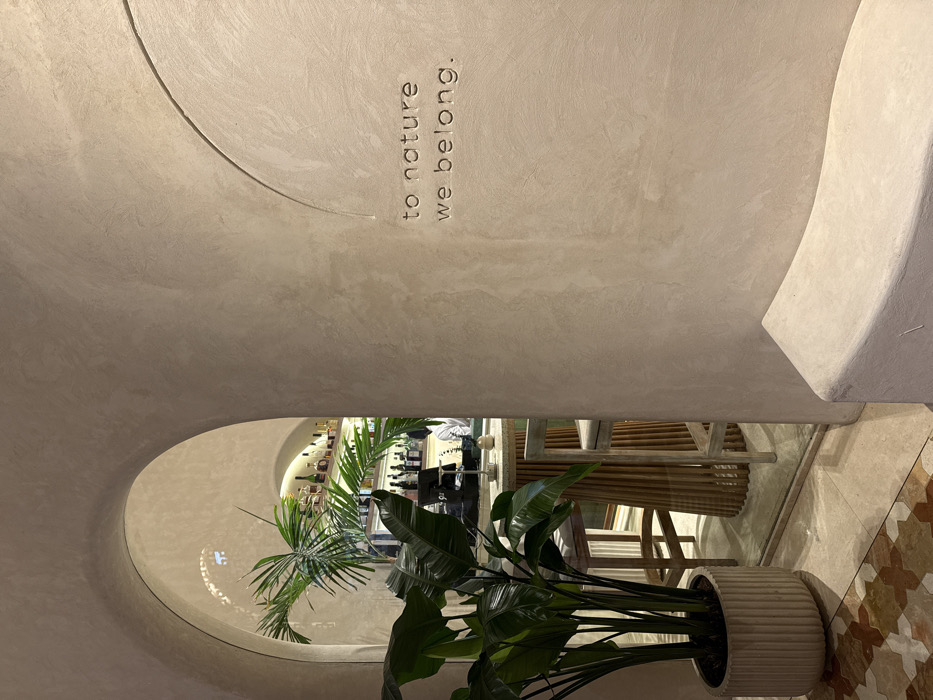 This image depicts an interior scene within an establishment located in Dubai, United Arab Emirates. The foreground features a light beige, textured wall with a prominent, curved arched opening on the left. On the right section of the wall, the phrase "to nature we belong." is etched in capital letters, with a subtle curved line appearing above it. A low, curved light-colored bench, matching the wall material, occupies the lower right corner. A large potted plant with broad, dark green leaves resides in a ribbed, off-white pot in the lower-left foreground. Through the arched opening, a bar area is visible. It features a round, light-colored bar counter with a base constructed from vertical wooden slats, surrounded by two light-framed bar stools with upholstered seats. Behind the counter, shelves display multiple bottles of varying shapes and colors. A person wearing a light-colored shirt is partially visible behind the bar, positioned to the right of a dark electronic device on the counter. Another plant, a smaller palm, is visible within the bar area. The floor in the immediate foreground features patterned tiles in cream, brown, and reddish hues, transitioning to a lighter, less detailed surface inside the bar.