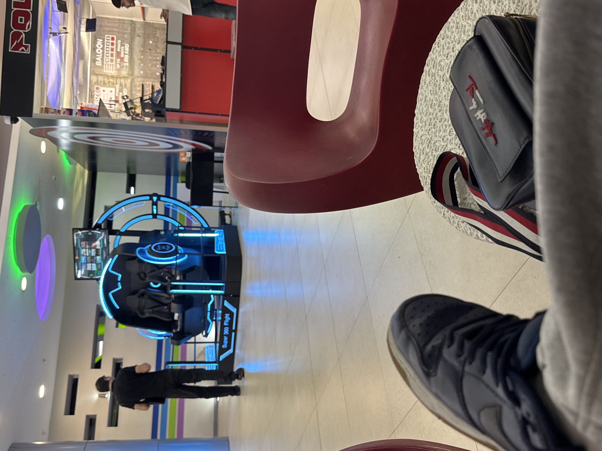 This image captures an interior scene within an entertainment center or arcade, likely in Yerevan, Armenia. The perspective is from a seated individual, with dark blue sneakers and light-colored pants partially visible in the bottom right. A red, ergonomically shaped chair is prominent in the mid-right. Resting on a white, textured surface next to the chair is a black bag with a prominent "Tommy Hilfiger" logo embroidered in red and white, and a striped strap (red, white, blue).
In the mid-ground, a person wearing a black t-shirt and black pants stands near a large, futuristic-looking virtual reality or simulation machine. This machine, labeled "Super 360 Flight," features intricate black structures with bright blue neon-like lighting accents, creating a dynamic appearance. A monitor displaying game options is visible above the machine.
The background shows more of the arcade environment. To the left of the VR machine, colorful circular light fixtures in green and purple adorn a wall. Further back, another arcade game is visible, with text that includes "BALOON," "SHOTS," and "UP TO POINTS 3,000 AMD," indicating a game that awards points or prizes in Armenian Dram (AMD). A partial view of another person is seen in the upper background. The floor is covered with light-colored tiles. The overall setting suggests an energetic indoor leisure space, likely during the daytime or evening, given the reliance on artificial lighting.
