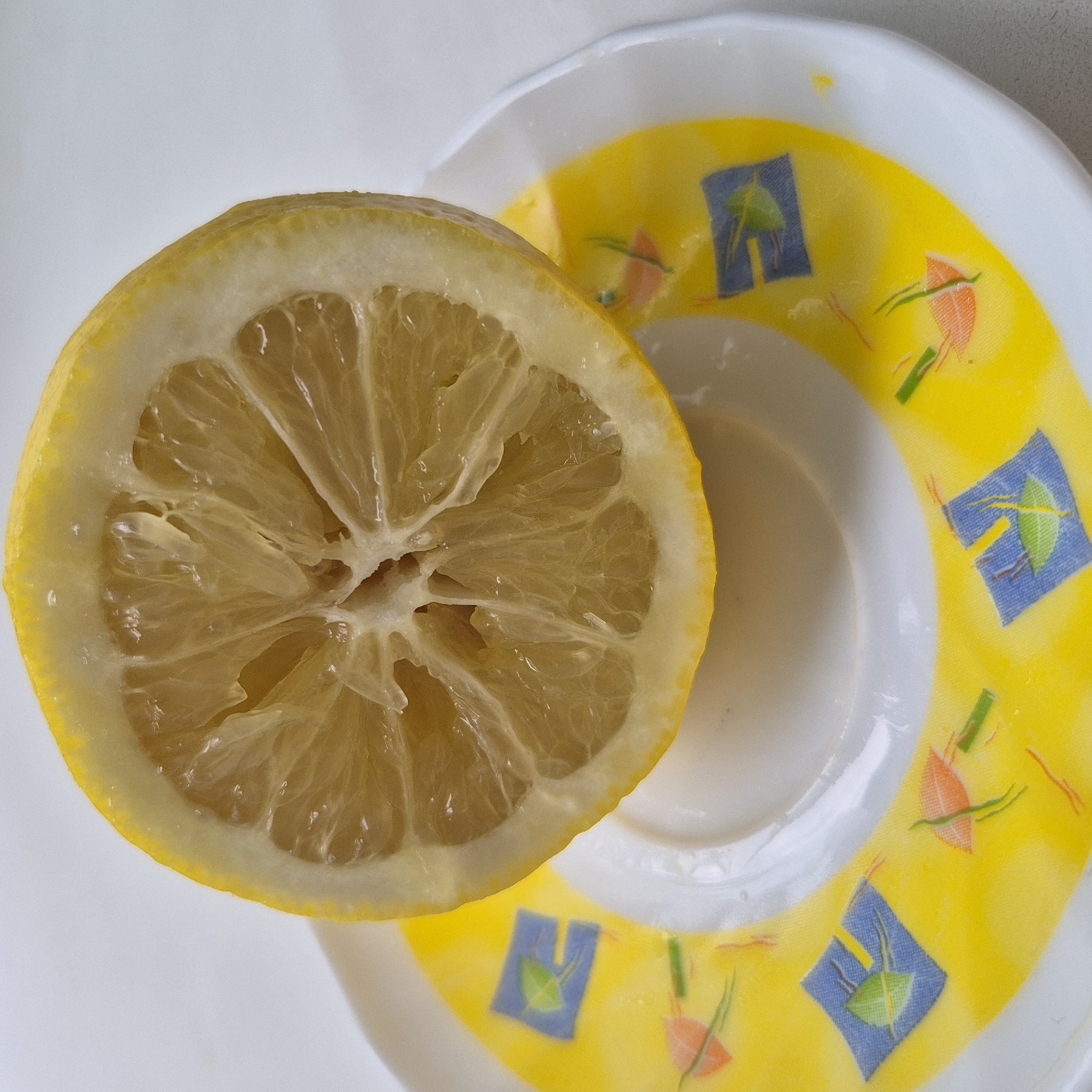 A close-up, top-down shot shows a half-slice of a bright yellow lemon resting on a white plate with a yellow rim. The lemon slice is positioned on the left side of the image, its textured flesh and pith clearly visible. The plate has a decorative pattern of colorful shapes, including blue squares with green designs and orange kite-like shapes with green lines, on its yellow rim. The background is a plain white surface. The lighting appears to be bright and even, suggesting an indoor setting. There is no visible text in the image. The scene evokes a sense of freshness and simplicity.