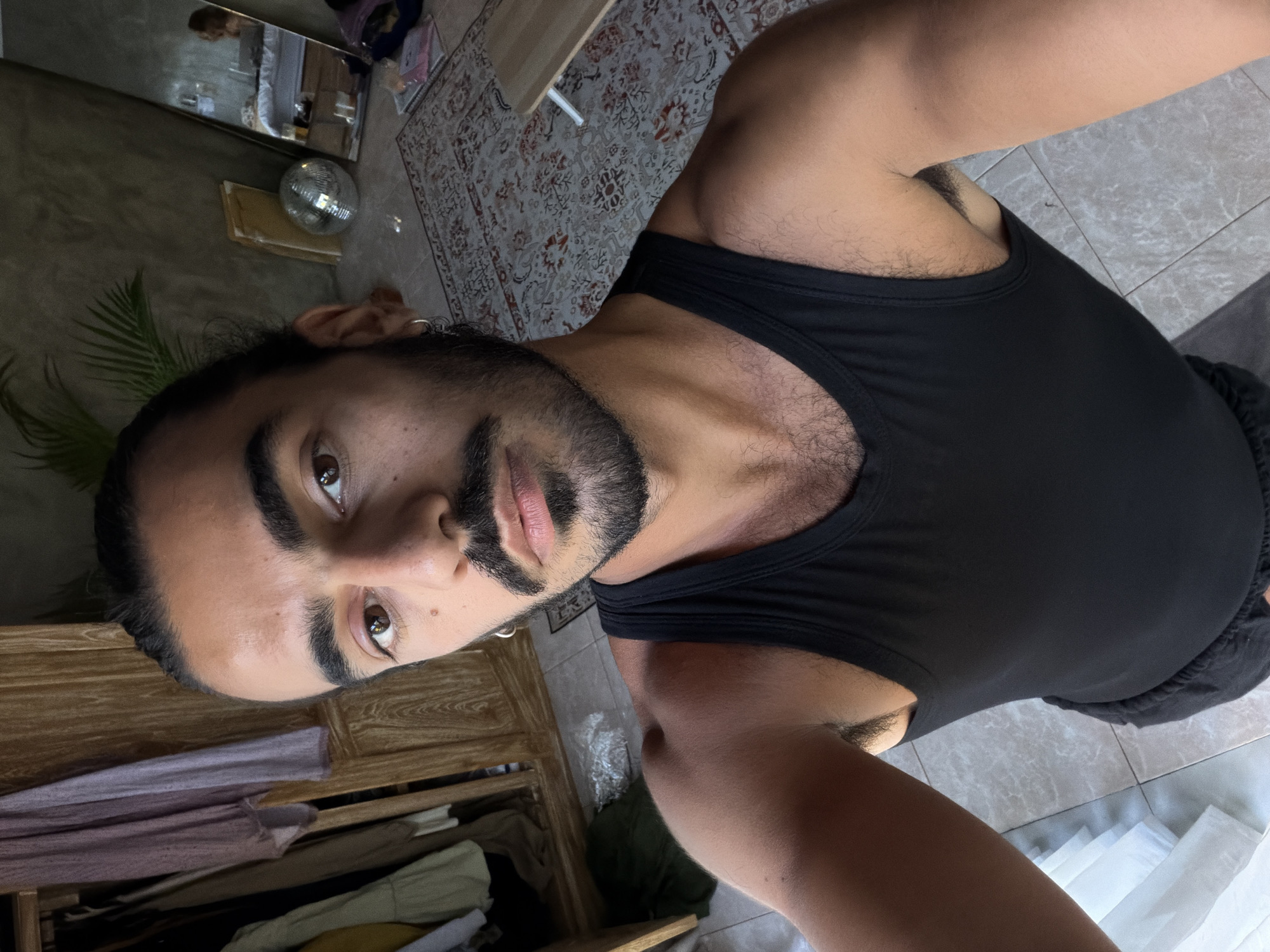 The image is a selfie taken by a person with medium-toned skin, dark hair pulled back into a bun, and a beard. They are wearing a black tank top and dark shorts. The person is standing indoors, in what appears to be a bedroom or living area. Behind them, a wooden wardrobe filled with clothes is visible. To their right, a large mirror reflects the room, showing a bed and a person sitting at a desk. A disco ball, a plant, and a patterned rug are also present in the room. The flooring is tiled. The location is identified as North Kuta, Indonesia.