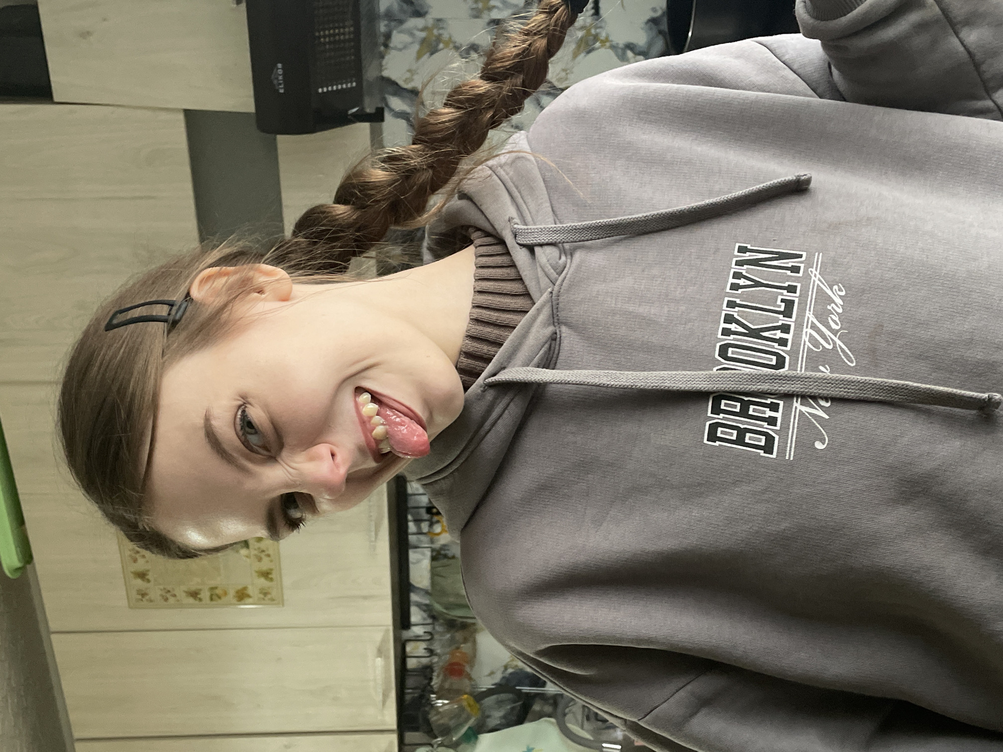 A playful portrait of a young person, likely female, captured indoors in a domestic setting, possibly a kitchen in Фоки, Russia. The image is taken from an unusual angle, rendering the subject upside down, which enhances the comedic and lighthearted nature of the photo.
The person has long, light brown hair styled into two distinct braids, one of which is visible cascading over her shoulder. A black barrette holds back some hair near her ear. She is wearing a grey hooded sweatshirt with a prominent drawstring and a brown ribbed collar visible underneath. The front of the hoodie displays white text: "BROOKLYN" in a bold, collegiate-style font, with "New York" written below it in an elegant cursive script.
Her facial expression is animated and humorous; she is sticking out her tongue, revealing her teeth, and her eyes are crinkled at the corners, suggesting a wide smile or laugh. Her eyes appear to be a light color.
The background includes light-colored wooden or laminate panels, indicative of cabinetry. A dark grey or black rectangular electronic device, which appears to be an extractor hood or air purifier, is mounted above the cabinetry. The brand "ELIKOR" is clearly visible on this device, and faint lettering suggests "RUS" might also be present, aligning with the Russian context of the brand. A patterned tile or decorative panel, resembling a kitchen backsplash, is seen on the wall behind her head. The lighting is artificial, consistent with an indoor setting, and no specific time of day is discernible. The overall impression is one of fun and spontaneity.