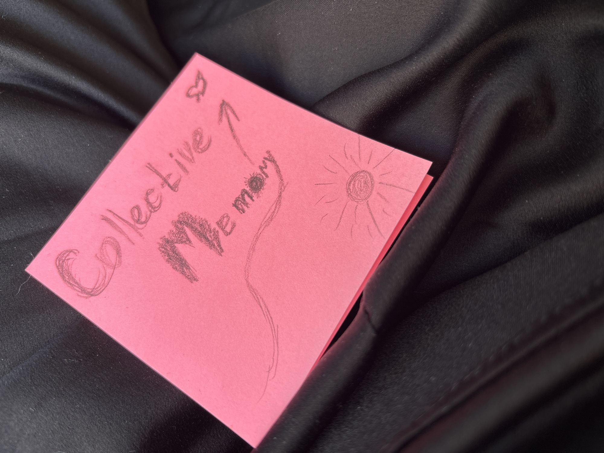 A pink sticky note sits on a wrinkled black fabric. The sticky note has the words "Collective Memory" written on it with a drawn heart, arrow, and sun. The location is unknown and there are no people shown in the image.