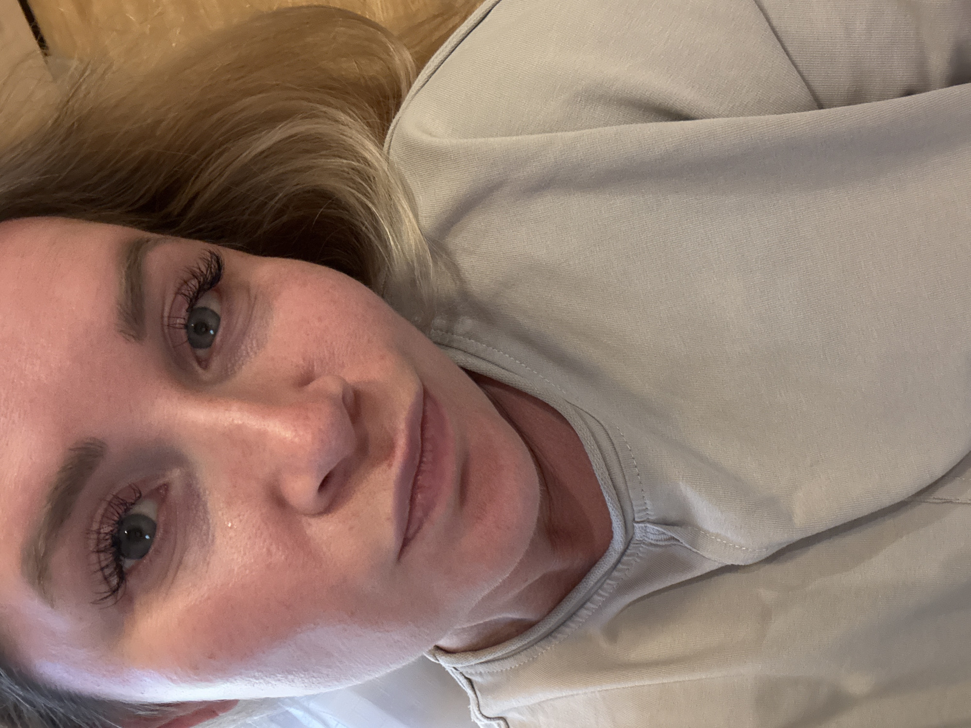 The image is a close-up selfie of a fair-skinned Caucasian woman with blonde hair. She is wearing a light beige crew-neck shirt. Her face is slightly flushed, and she appears to have makeup on, including mascara and possibly false eyelashes. Her eyes are light blue or green. The background is a wooden surface, suggesting an indoor setting. The image does not provide any information about the city of Veliky Novgorod, Russia, or any specific location within it. There are no visible objects or activities that would indicate a particular setting or context.