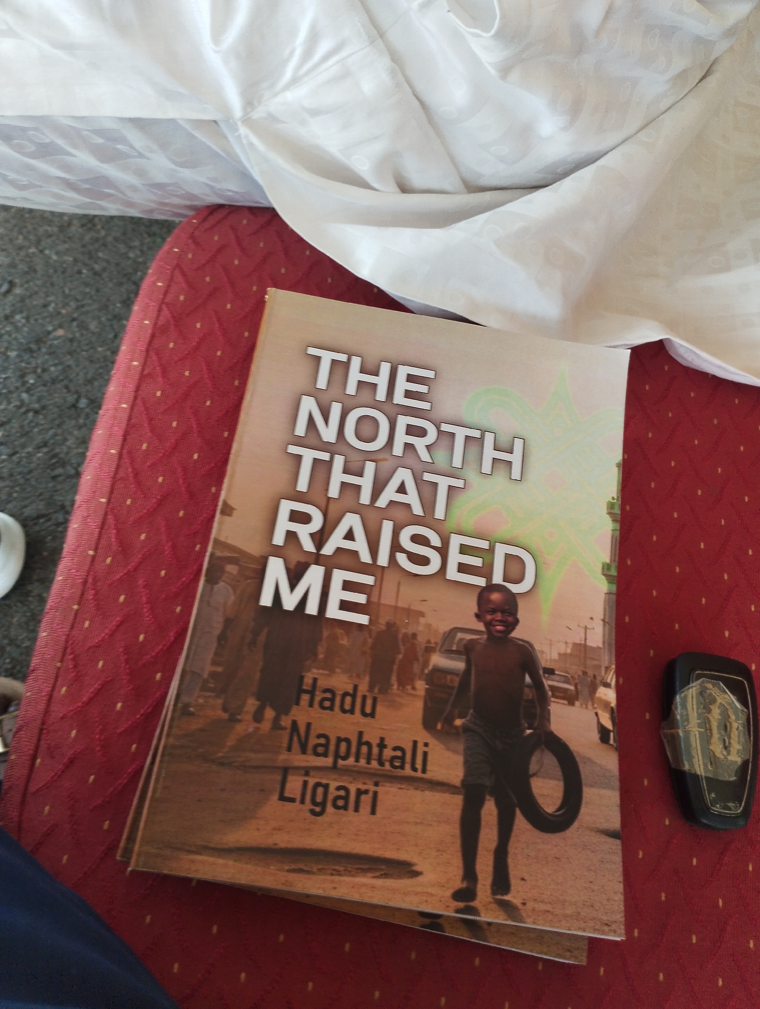 The image shows a book titled "The North That Raised Me" by Hudu Naphtali Ligari, resting on a red patterned surface, likely a seat. The book cover features a photograph of a young, smiling, dark-skinned boy carrying a tire in a dusty urban environment. In the background, there are blurred figures of people, cars, and buildings, indicative of a street scene. The photograph on the book is of the city of Gombe, Nigeria. A black object, possibly a key fob, lies next to the book on the right. Draped above the scene is a white cloth.