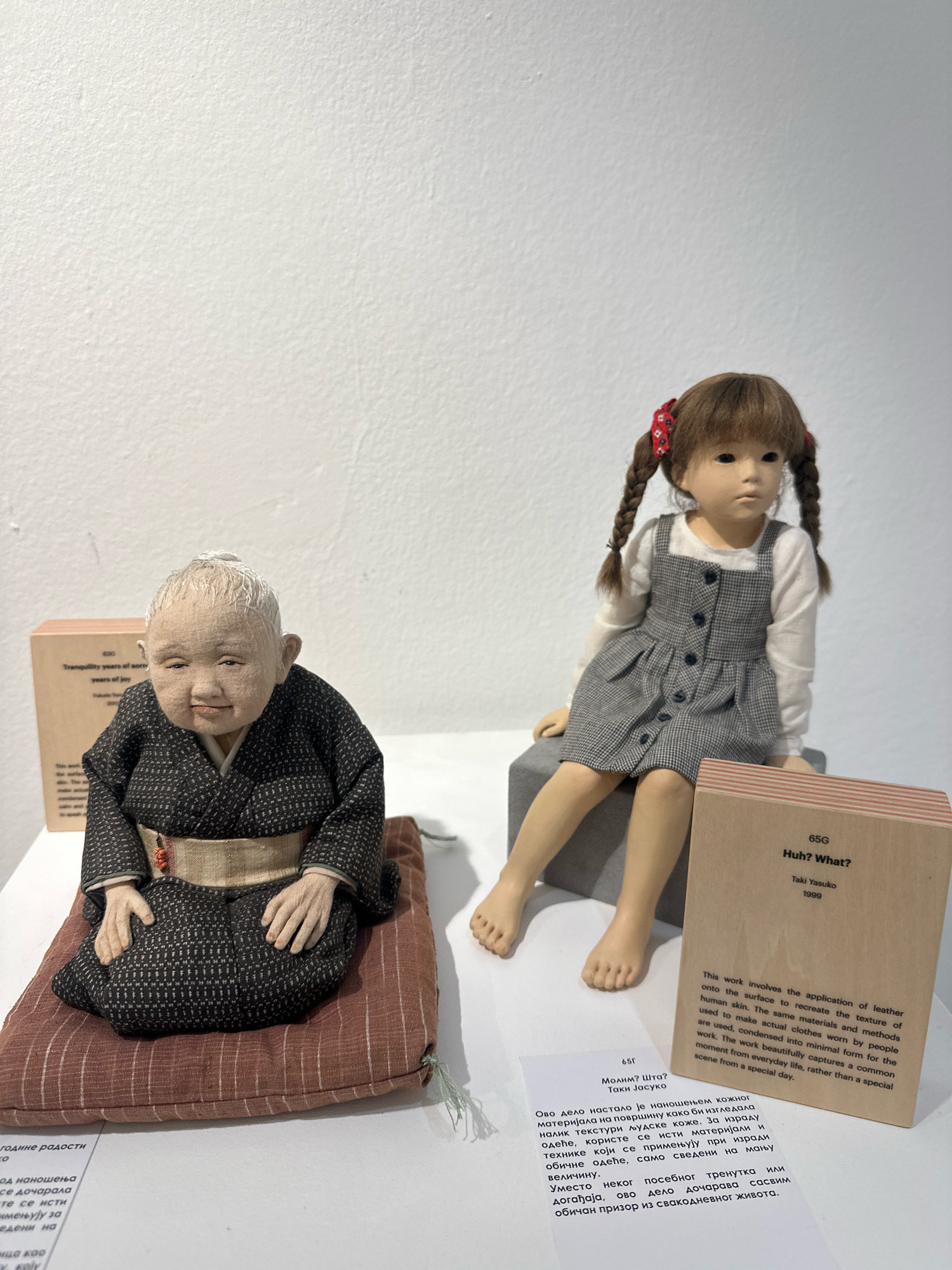 The image shows a display at the Museum of Contemporary Art in Belgrade, Serbia. On the left, a lifelike doll of an elderly person in traditional Japanese attire sits cross-legged on a patterned cushion. To the right, a doll of a young girl with braided hair, wearing a gray and white checkered dress over a white top, is seated on a gray block. In front of the dolls are two display cards with text. The card in front of the elderly doll is partially visible and appears to have text in Serbian. The card in front of the young girl's doll has English text and reads "65G Huh? What? Taki Yasuko 1999". Both dolls are rendered with intricate detail, showcasing realistic skin texture and clothing. The background is a plain white wall, suggesting an indoor exhibition space.