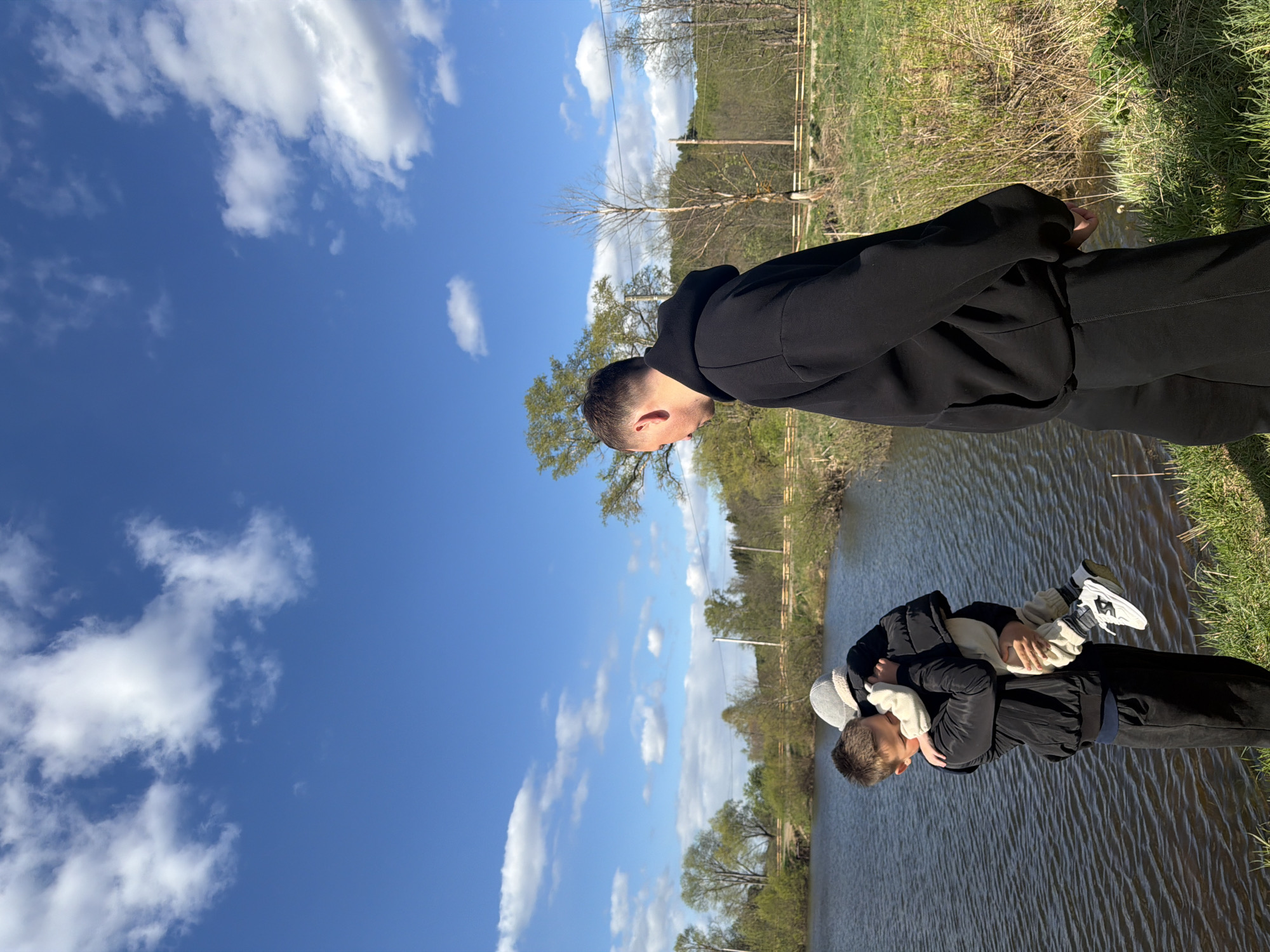 A man in a black hooded sweatshirt and pants stands by a body of water, presumably in Большой Букор, Russia. He holds a small child dressed in a jacket, hat, and shoes with white soles. The sky is blue with scattered white clouds. The background includes trees, foliage, and what appears to be a metal railing or fence.