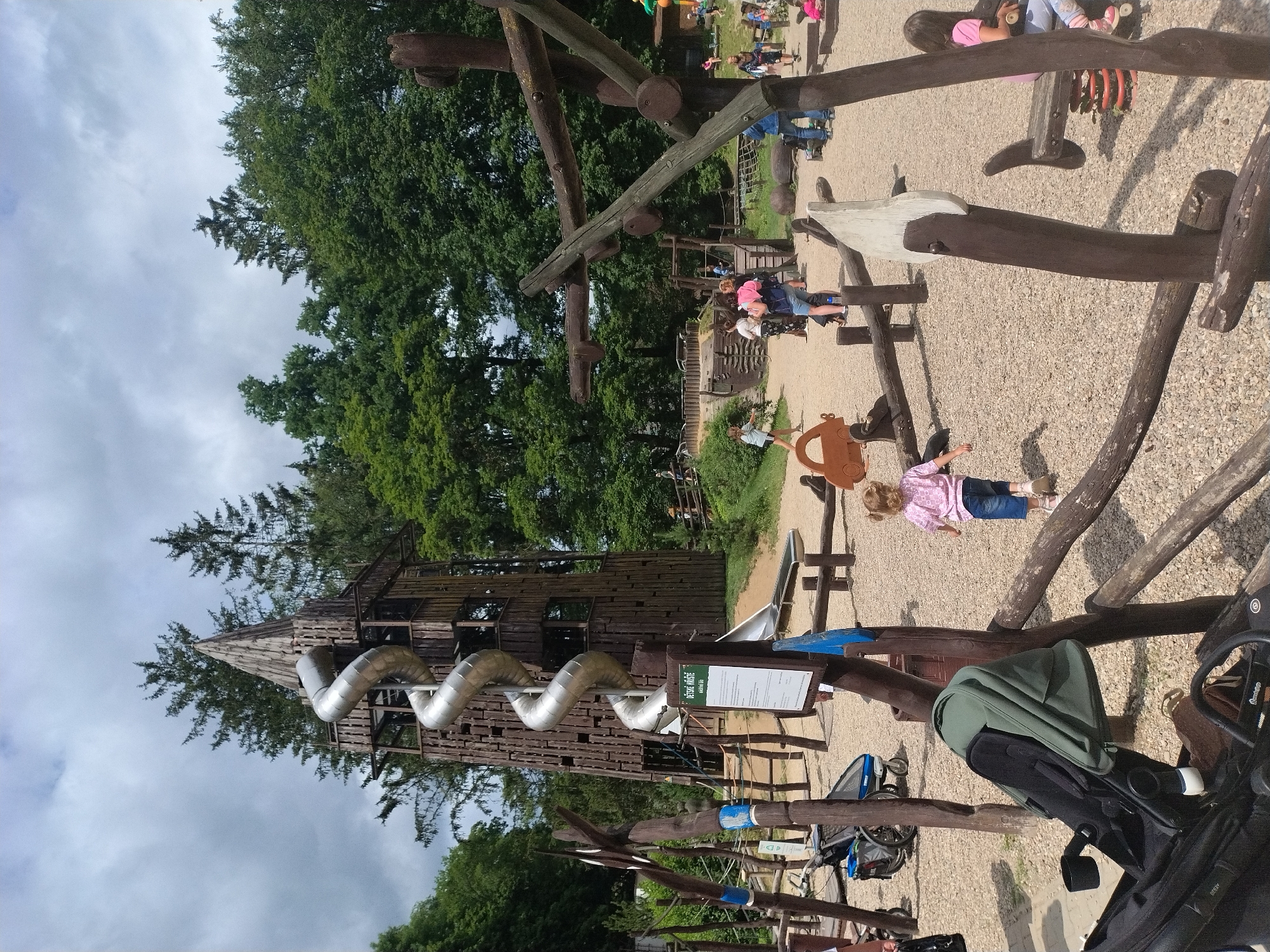The image depicts an outdoor playground in Jihlava, Czechia, characterized by natural materials like wood and gravel. A prominent wooden tower with a spiral metal slide dominates the background, surrounded by lush green trees under a cloudy sky. In the foreground and midground, various wooden play structures are visible, including a seesaw, a rocking horse, and low wooden fences. Several people are present, including children playing and adults supervising. One child in a pink shirt and denim shorts is running away from the camera. A stroller is parked on the left side of the frame. The ground is covered with small stones. A sign with Czech text is attached to a wooden post.