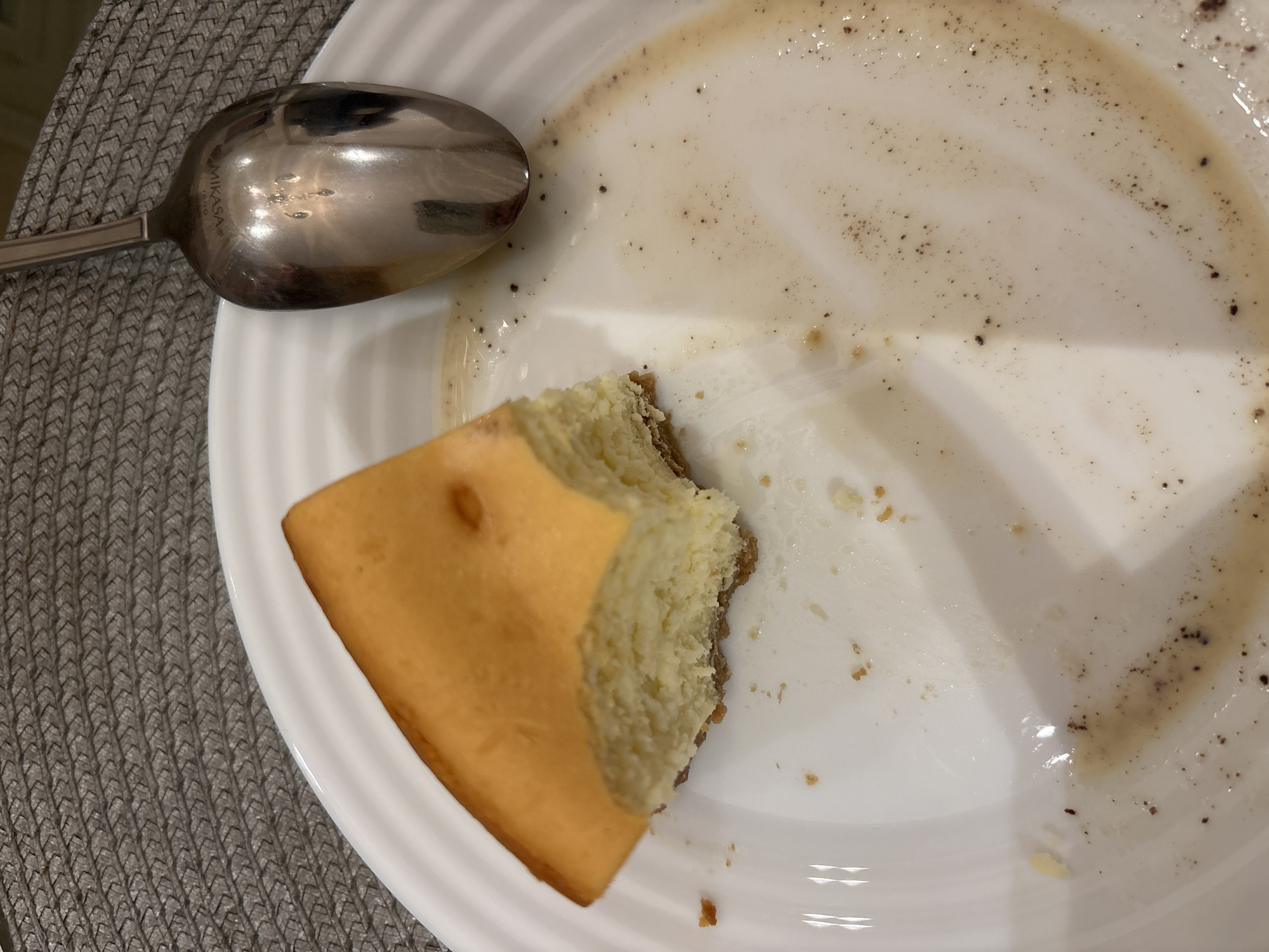 In Mexico City, Mexico, a partially consumed slice of cheesecake rests on a white ceramic plate with a ridged rim. The cheesecake exhibits a golden-brown top crust, a creamy yellow interior, and a visible bite mark on its left side. A silver-toned metal spoon, engraved with "MIKASA" and "18/10", lies on the upper right portion of the plate. The plate surface is marked with cheesecake crumbs, moisture, and a light brown, circular stain interspersed with small dark specks. The plate and spoon are positioned on a dark grey, woven placemat. No individuals are present in the scene; the items indicate recent consumption.