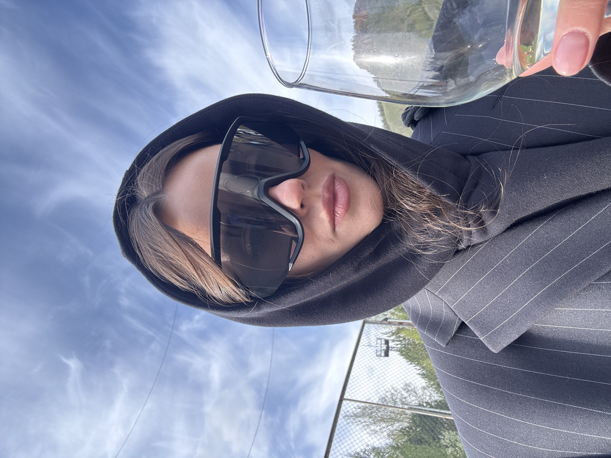 A woman wearing a dark pinstripe blazer and a dark hooded sweatshirt is holding a partially full glass of water. She is wearing large sunglasses that reflect the outdoors. The background shows a blue sky with white clouds, a chain-link fence, and some greenery. The image appears to have been taken outdoors in daylight. There is no explicit indication of the location being Krasnoyarsk, Russia, in the visible details of the image.