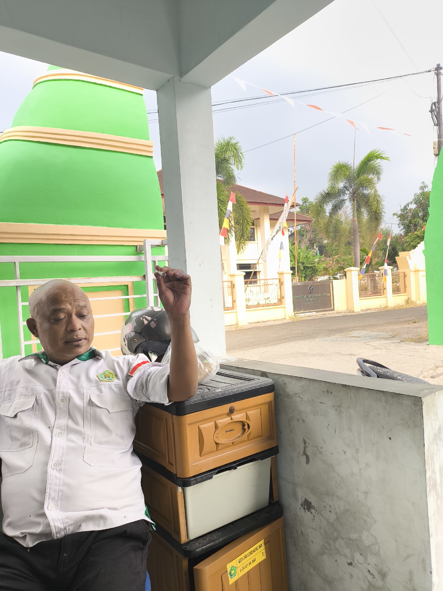 The image shows a man sitting outdoors, holding a cigarette. He is wearing a white shirt with a green emblem on the left chest. Behind him, there are stacks of plastic storage drawers. In the background, there is a building with a sign that reads "SDN PONDOK CABANG BALONG" and a wall with an inscription. Several flags are visible, including Indonesian flags. The man is bald and appears to be middle-aged. The scene appears to be in Cilegon, Indonesia, based on the building sign. The overall environment is an outdoor area, likely a public space or school grounds.