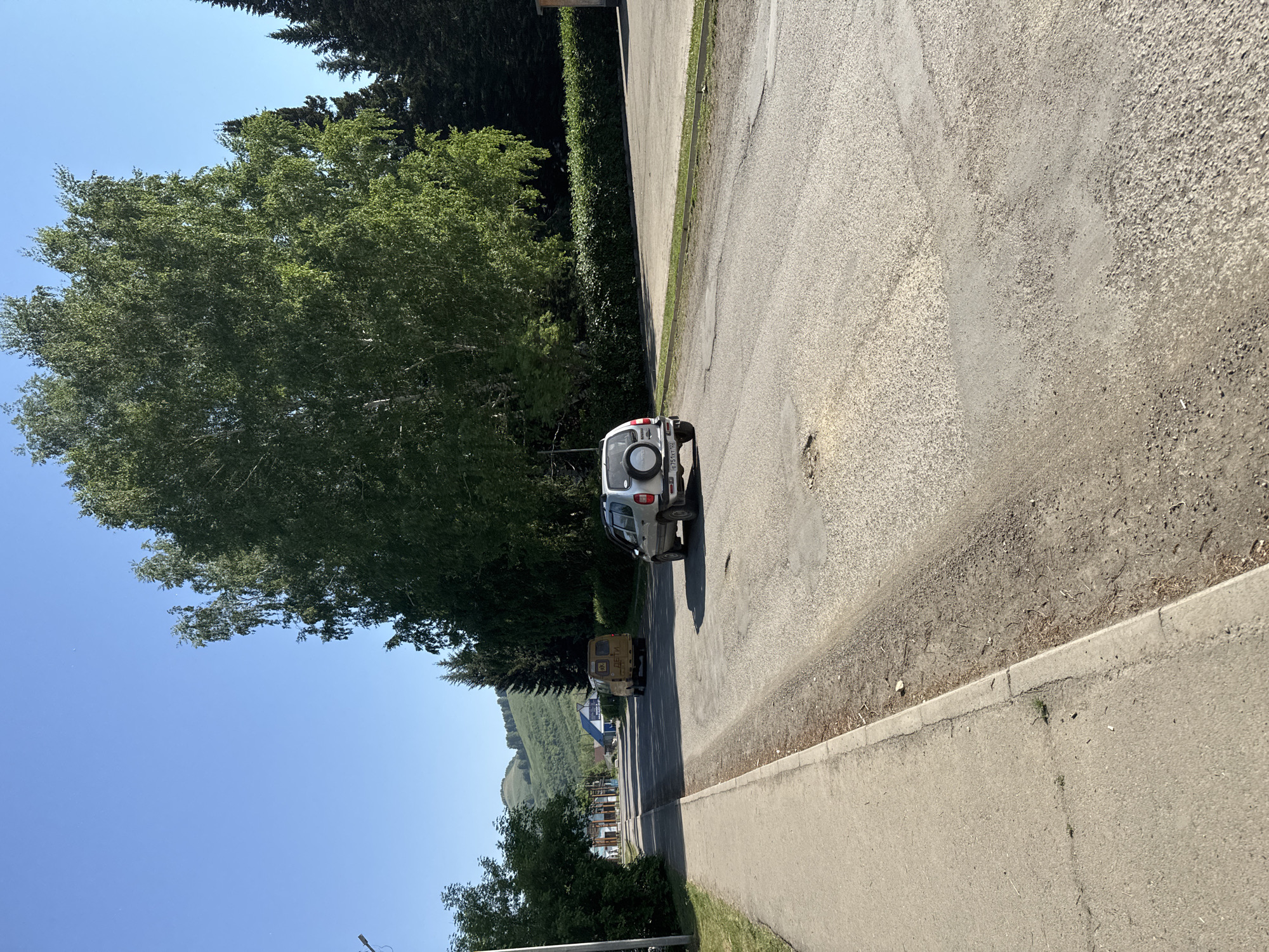 In the town of Soloneshnoye, Russia, on a bright, sunny day, a paved road runs through a semi-rural area. The road itself is in poor condition, with visible potholes and patched sections. On the right side of the road, a silver SUV with a spare tire mounted on its rear door is driving away from the camera. On the left side of the road, a sidewalk runs alongside the pavement, bordered by dry grass and dirt. Further down the road, a yellow van is also visible, and in the distance, buildings and a tree-covered hillside can be seen. Large, leafy trees line the right side of the road, with a dense hedge in front of them. The sky is a clear, vibrant blue.