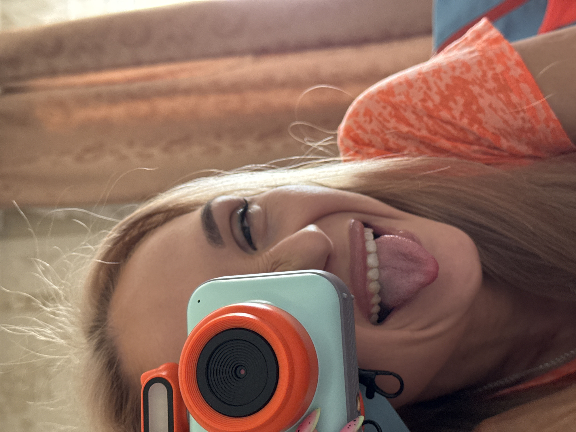 A close-up, rotated selfie captures a young woman lying down and making a playful expression, sticking out her tongue and squinting one eye. Her long, light-colored hair, appearing somewhat windswept, frames her face. She has defined eyebrows and a broad, open-mouthed smile, revealing her teeth. In the foreground, partially obscuring the lower left of her face, is a vibrant light blue and orange digital camera, likely a toy or novelty item, featuring a prominent orange lens ring and a small orange side handle. She is dressed in an orange top with a subtle, lighter-toned pattern, with a hint of a blue garment visible near her shoulder. The background is softly blurred with warm, light brown and beige hues, suggesting an indoor setting such as curtains or bedding. The overall mood is humorous and lighthearted. No text is visible in the image. The time of day and weather conditions are not discernible. The location context provided is Chaikovsky, Russia.