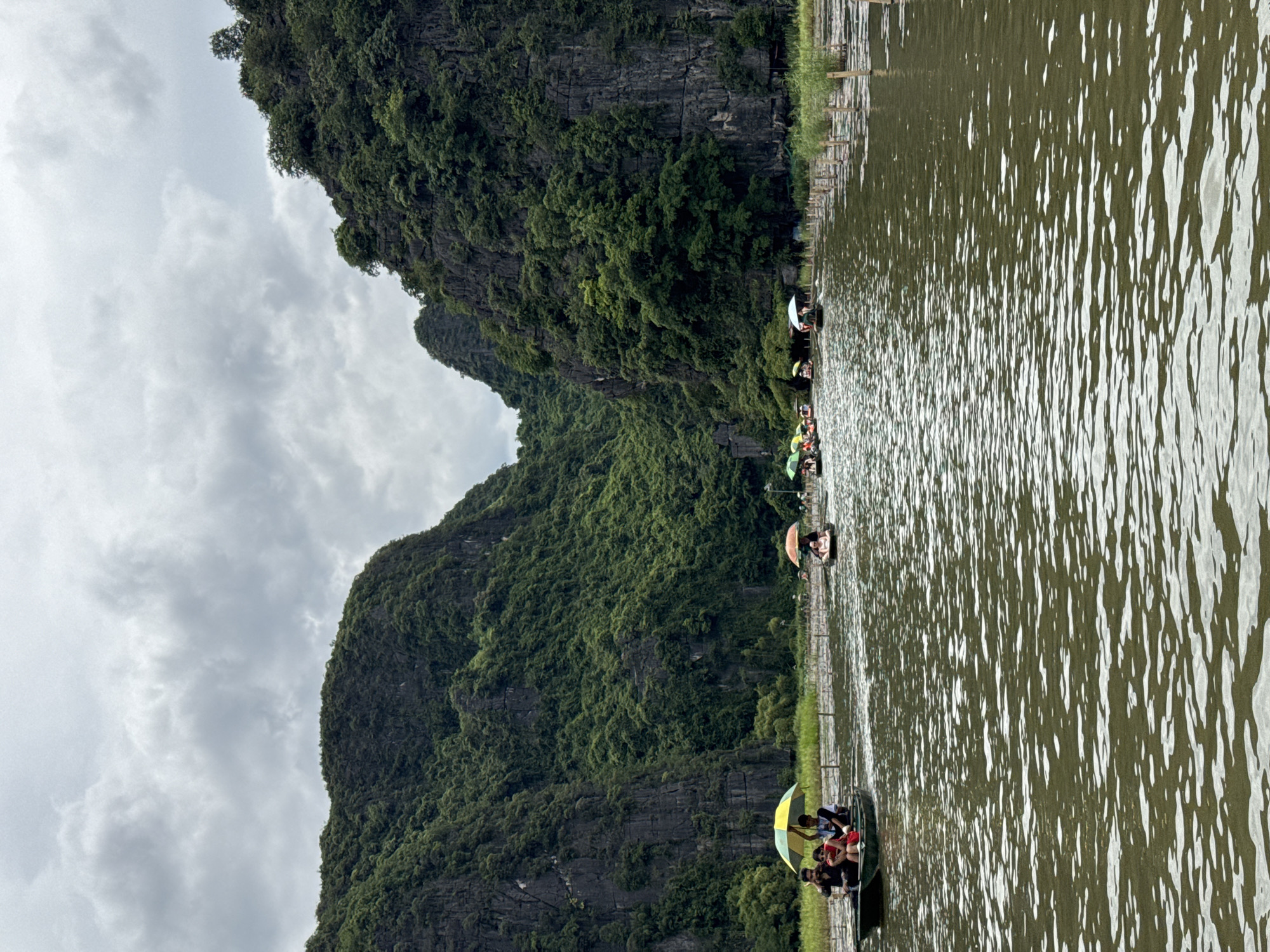 The image shows a body of water with several small boats carrying people. The boats are navigating through a narrow channel between two large, steep limestone mountains covered in lush green vegetation. The sky is overcast with gray clouds. In the foreground, the water is rippled, reflecting the muted light. Several boats are visible, some with people holding colorful umbrellas for shade. One boat in the foreground has three people, two seated and one standing, with a yellow and green umbrella. Further in the distance, more boats with people and umbrellas can be seen. The mountains rise dramatically on either side, creating a sense of awe and natural grandeur. Along the right bank of the waterway, a series of wooden poles are sticking out of the water, possibly marking a path or part of a farming/fishing structure. The overall scene suggests a popular tourist activity in a scenic natural environment, likely a river or lake tour. The location is identified as Nam Hoa Lư Ward, Vietnam.