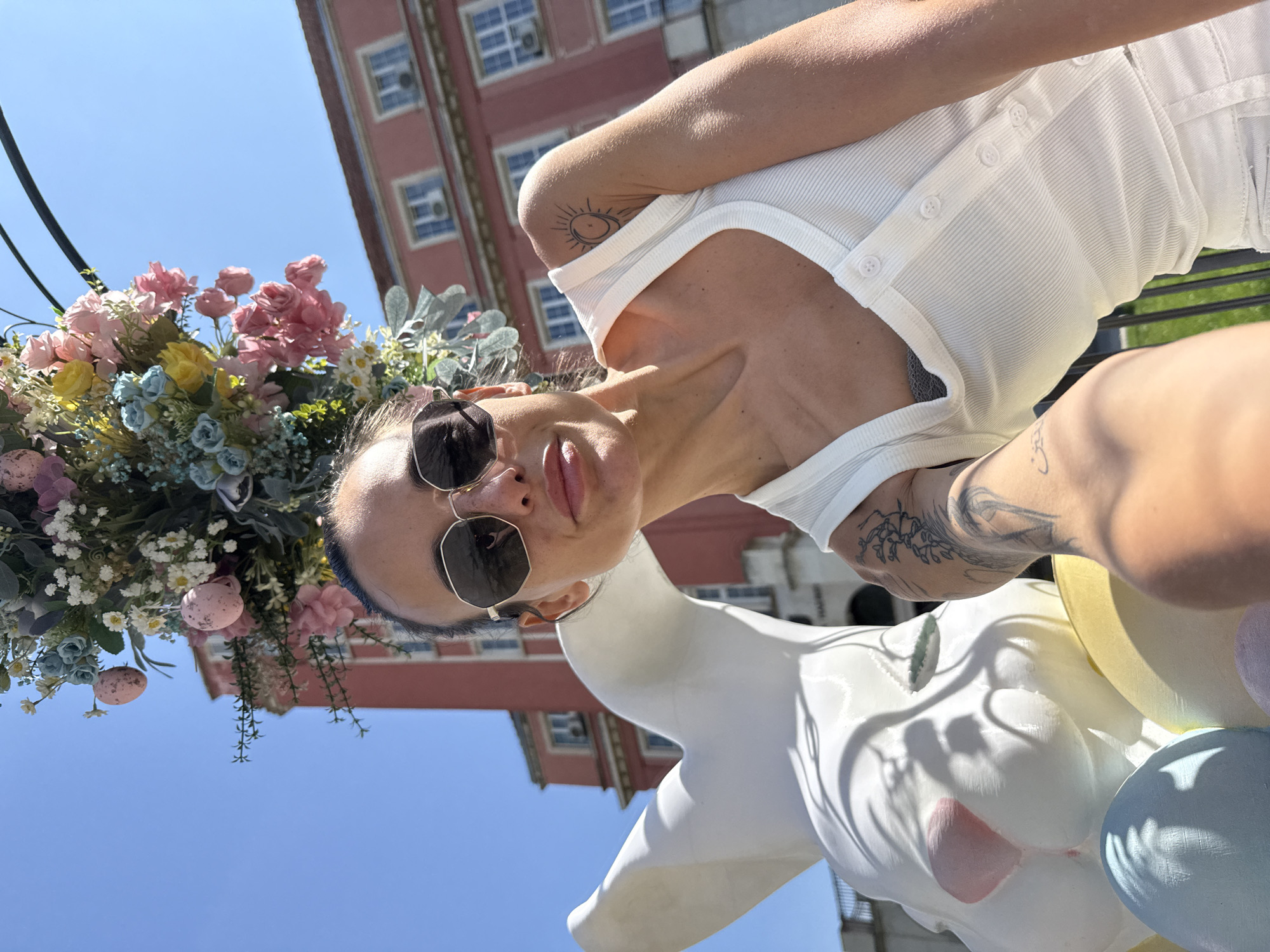 A woman takes a selfie with the 'Plevunka' sculpture in Pleven, Bulgaria, on a sunny day. The woman wears sunglasses and a white top. The sculpture is white with pastel-colored accents. Flowers and decorative eggs adorn the top of the sculpture. In the background, a red building with windows is visible against a blue sky.