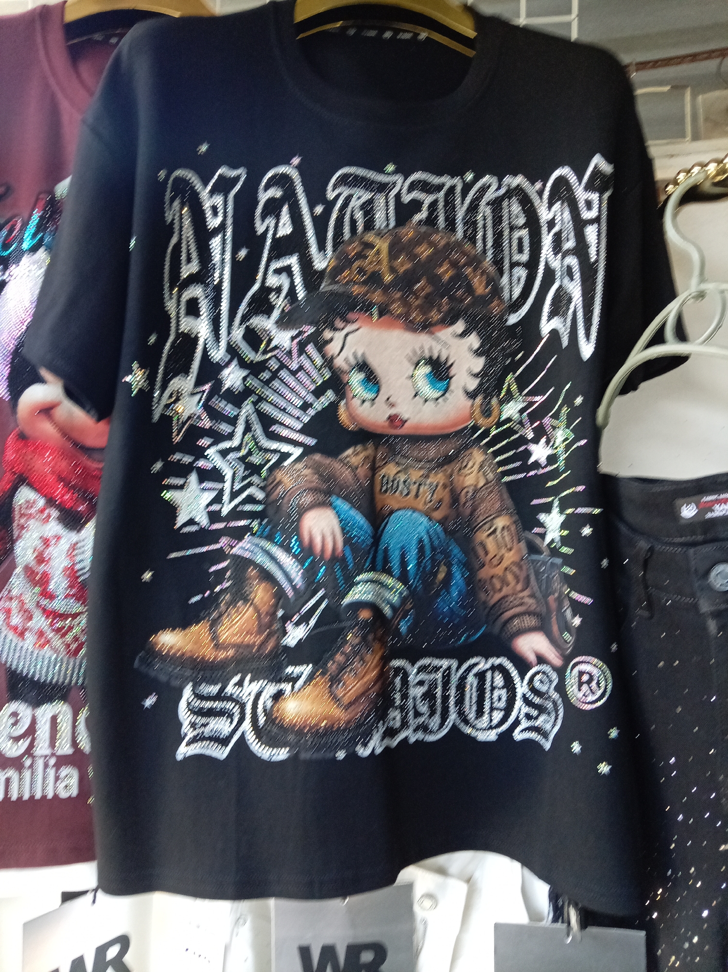 A black t-shirt featuring a large graphic of Betty Boop with embellishments is displayed. The graphic shows Betty Boop wearing a bejeweled baseball cap and a sweater with the word "BOOSTY" in rhinestones. She is depicted sitting down with blue jeans and brown boots, surrounded by stars and sparkly accents. The t-shirt is hanging on a clothing rack, with another t-shirt in a muted red color visible behind it. The scene appears to be in a retail setting, possibly a store, with other garments and price tags partially visible. The image does not provide any information about the city of Donetsk, Ukraine.