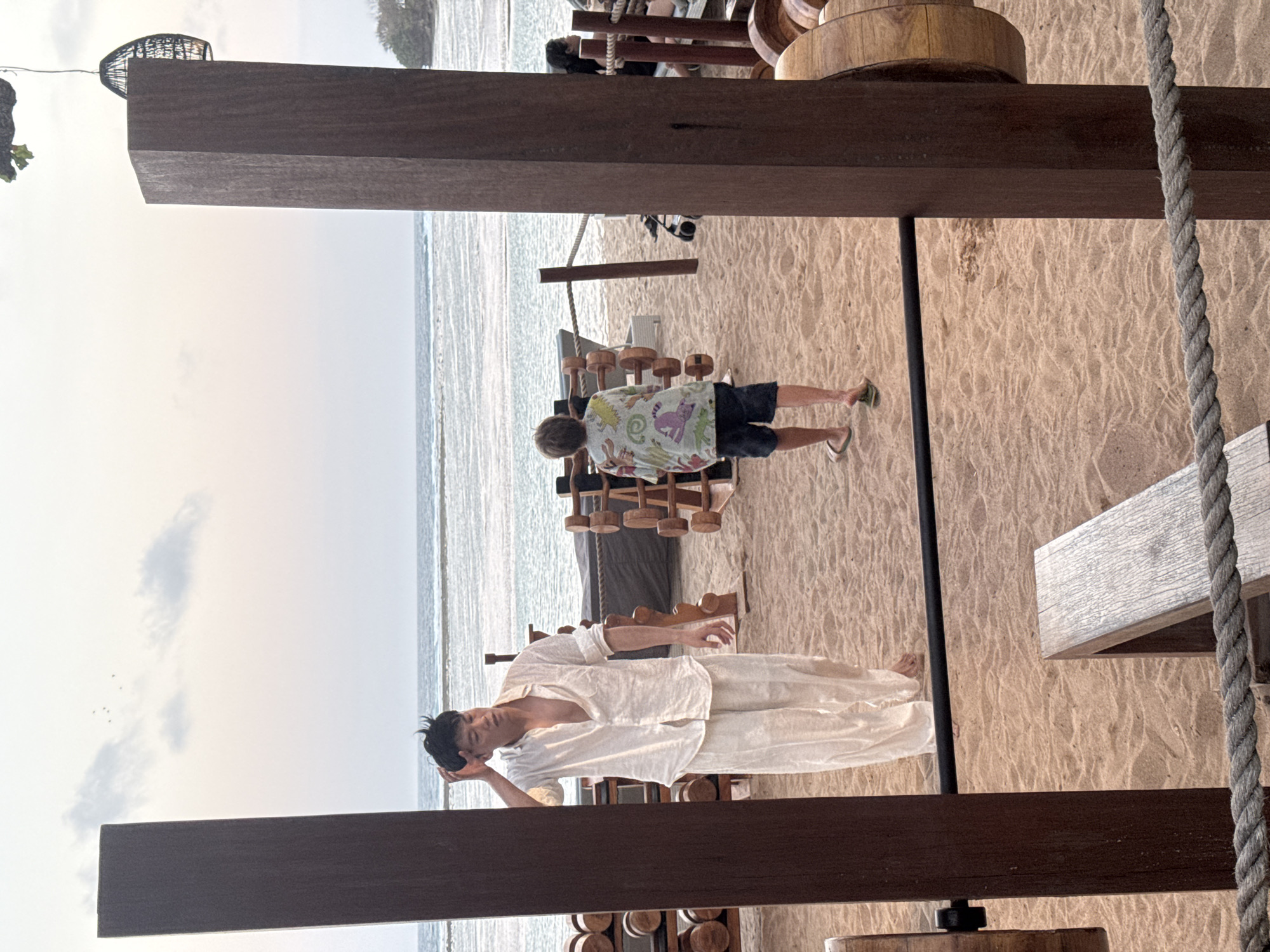 In Kuta Selatan, Indonesia, a beachside gym features two individuals. On the left, a man in loose-fitting white clothing stands with his hand on his head, facing away from the ocean. To his right, a younger person in a patterned shirt and dark shorts walks towards the water, also with their back to the viewer. The gym equipment, made of wood and metal, is arranged on the sand. In the background, the ocean stretches to the horizon under a cloudy sky, with gentle waves lapping the shore.