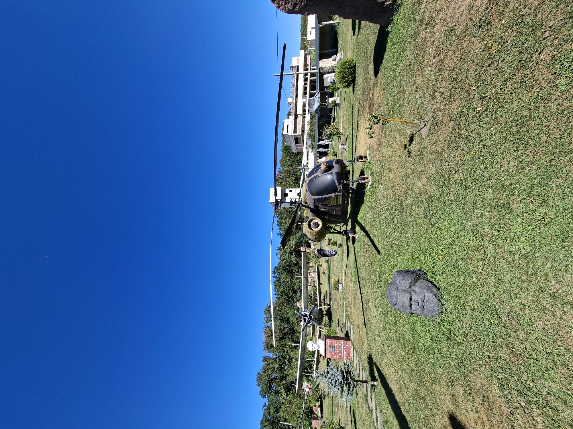 In Malcoci, Moldova, a sunny outdoor scene features several historical aircraft and a helicopter displayed on a grassy lawn. In the foreground, a large, dark stone sculpture of a human head with a beard and long hair rests on the grass. Behind it, a dark green military helicopter is positioned. Further back, a white biplane is visible with a bust of a person on a brick pedestal next to it. In the distance, a multi-story building with a modern architectural style stands amidst trees, under a clear blue sky. The environment appears to be a park or an open-air museum.