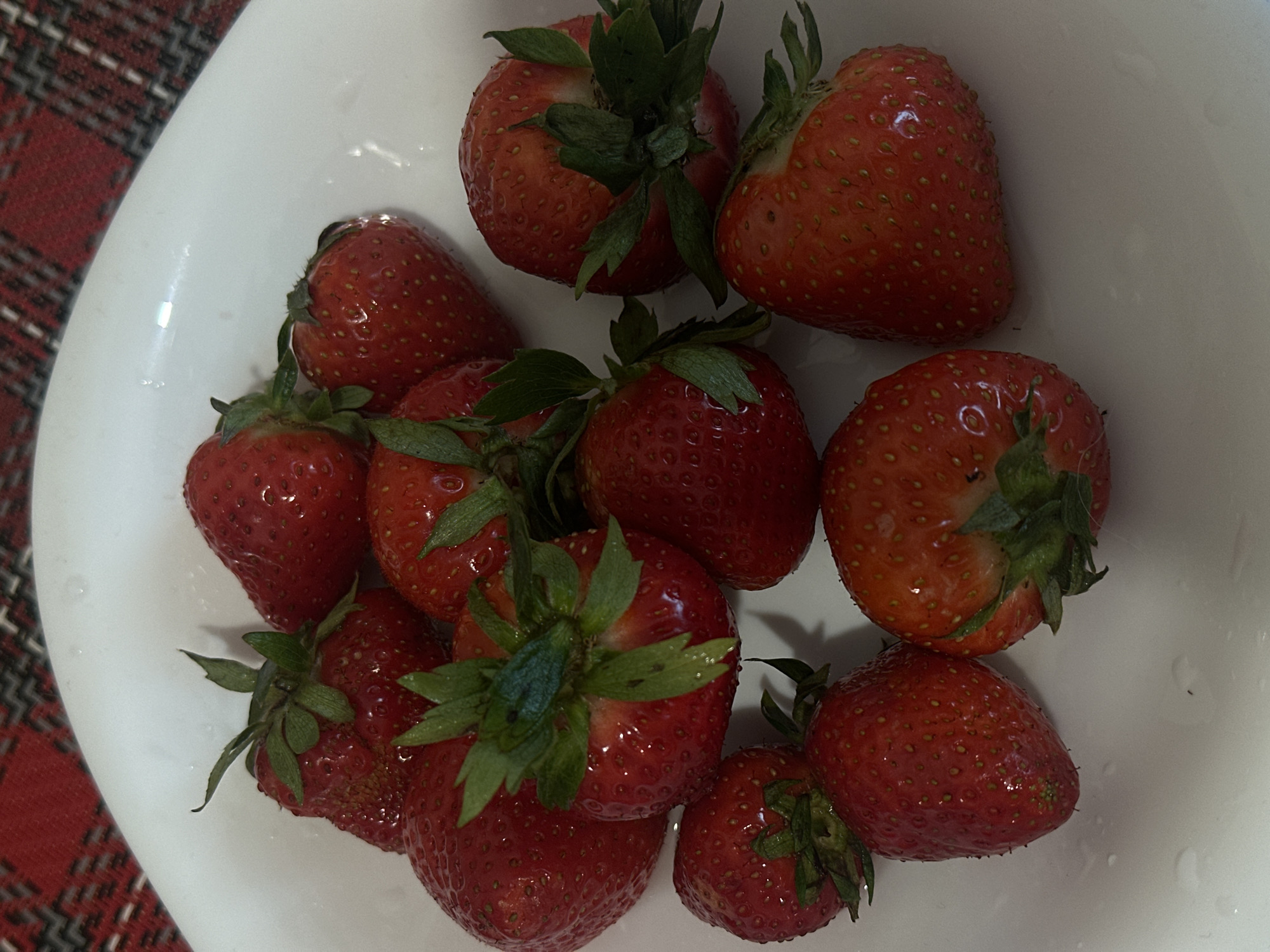 A top-down view shows a white bowl containing approximately ten fresh, red strawberries with green leafy tops. The bowl is positioned on a surface covered with a red and black plaid pattern. The strawberries are shiny, likely due to being washed. No people are visible in the image.
This image does not contain information that would allow for determination of the location in the city of Votkinsk, Russia.